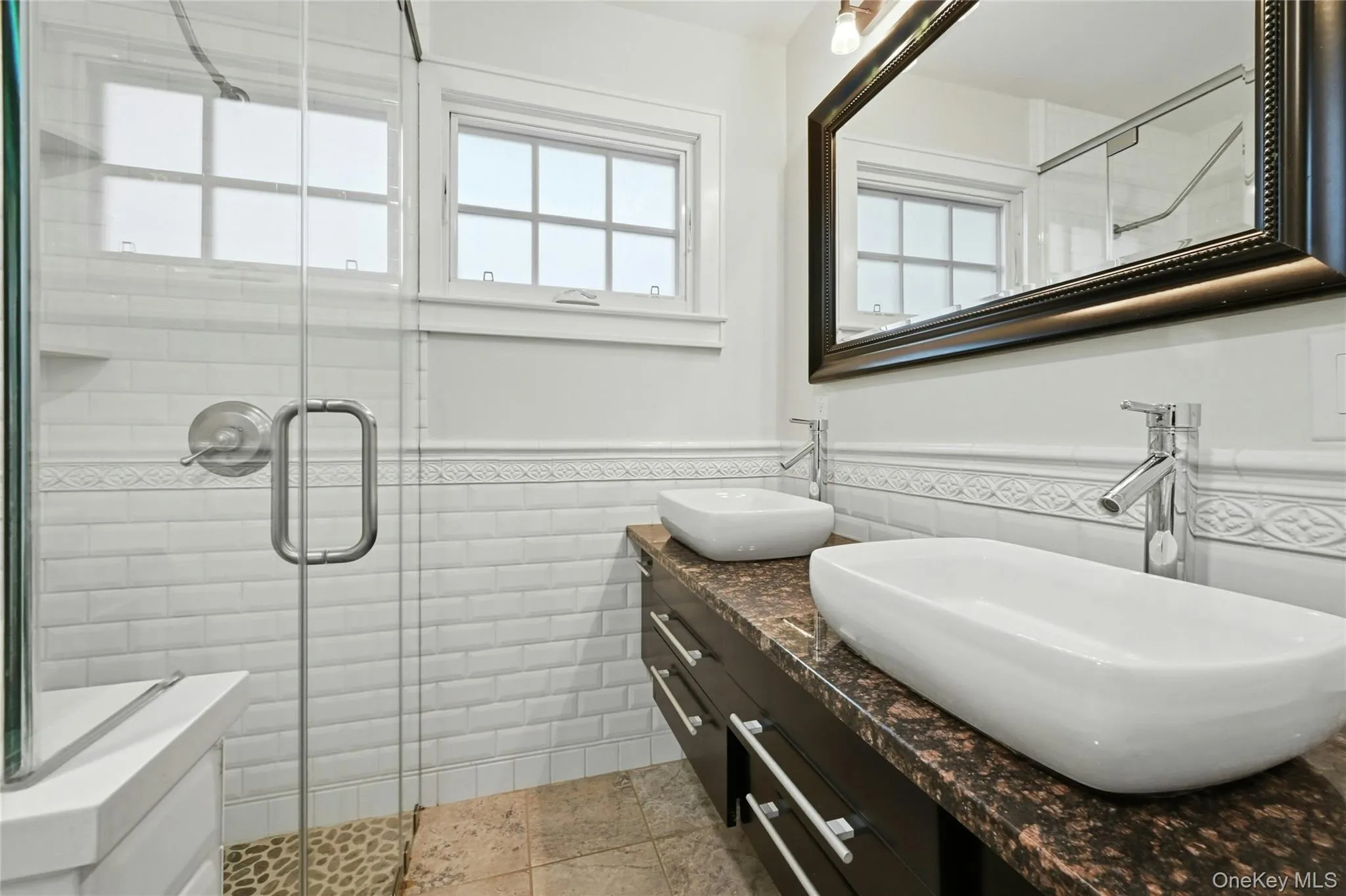Full bath featuring double vanity, wainscoting, tile walls, healthy amount of natural light, and a stall shower Full bath featuring double vanity, wainscoting, tile walls, healthy amount of natural light, and a stall shower
