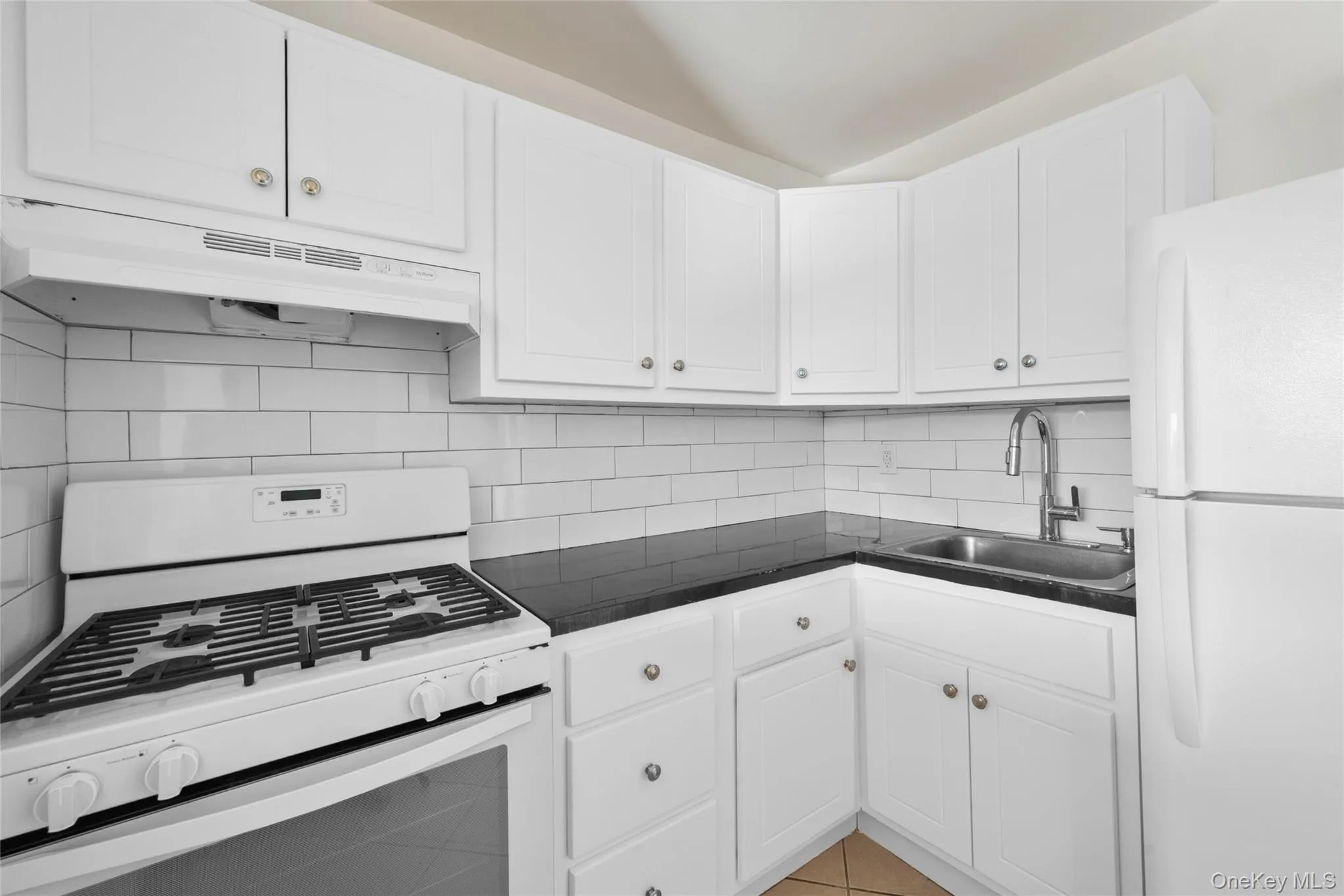 Kitchen with white appliances, dark countertops, white cabinets, under cabinet range hood, and backsplash Kitchen with white appliances, dark countertops, white cabinets, under cabinet range hood, and backsplash