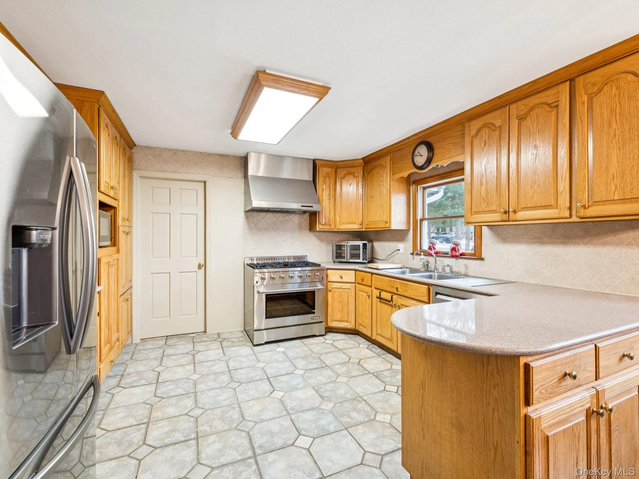 Kitchen featuring stainless steel appliances, wall chimney range hood, brown cabinets, light stone counters, and light flooring Kitchen featuring stainless steel appliances, wall chimney range hood, brown cabinets, light stone counters, and light flooring