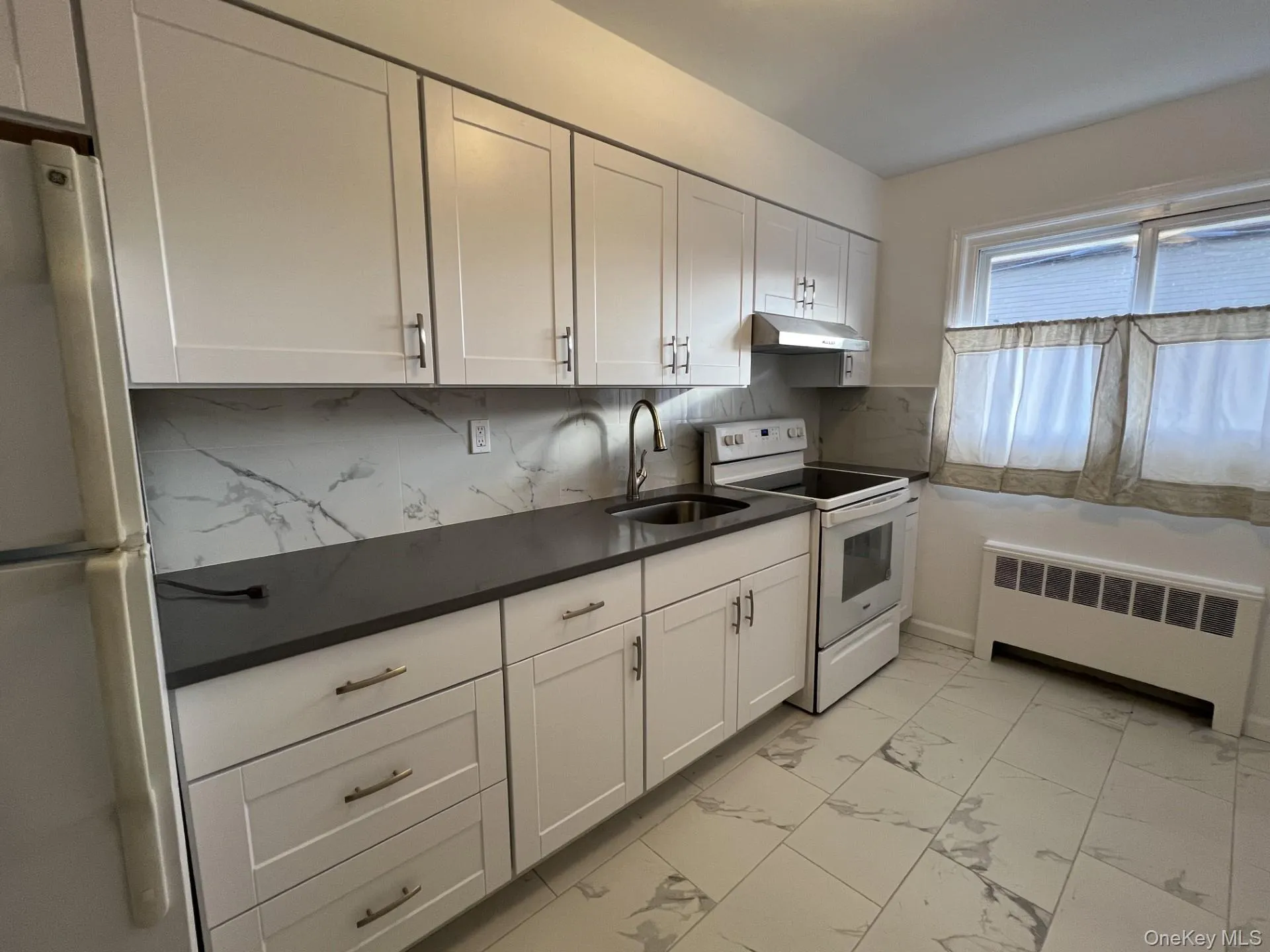 Kitchen with white appliances, radiator heating unit, white cabinets, tasteful backsplash, and light marble finish floors Kitchen with white appliances, radiator heating unit, white cabinets, tasteful backsplash, and light marble finish floors