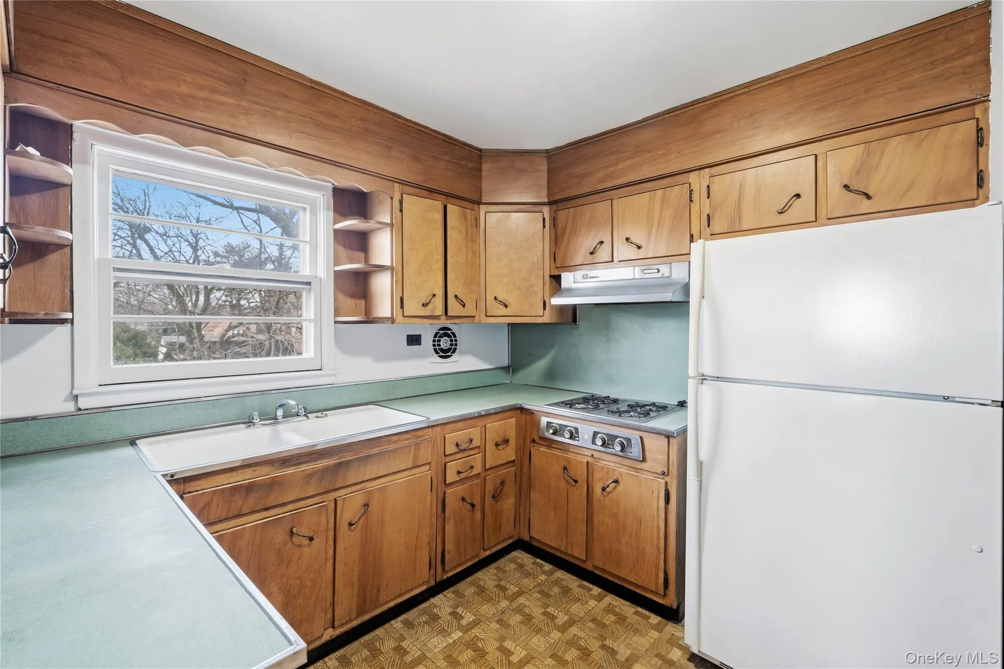 Kitchen with open shelves, freestanding refrigerator, light countertops, and brown cabinetry Kitchen with open shelves, freestanding refrigerator, light countertops, and brown cabinetry