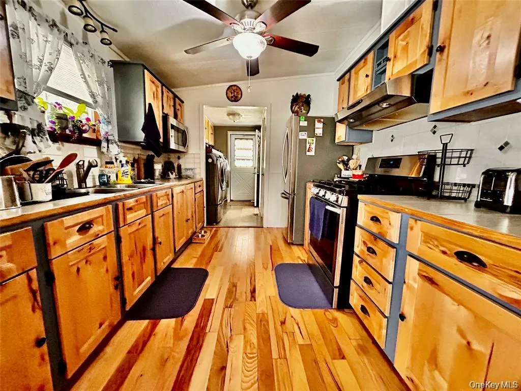 Kitchen featuring stainless steel appliances, under cabinet range hood, light wood-style floors, brown cabinetry, and ceiling fan Kitchen featuring stainless steel appliances, under cabinet range hood, light wood-style floors, brown cabinetry, and ceiling fan