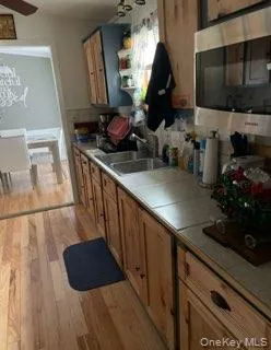 Kitchen with tile countertops, stainless steel microwave, light wood-style flooring, a ceiling fan, and brown cabinetry Kitchen with tile countertops, stainless steel microwave, light wood-style flooring, a ceiling fan, and brown cabinetry