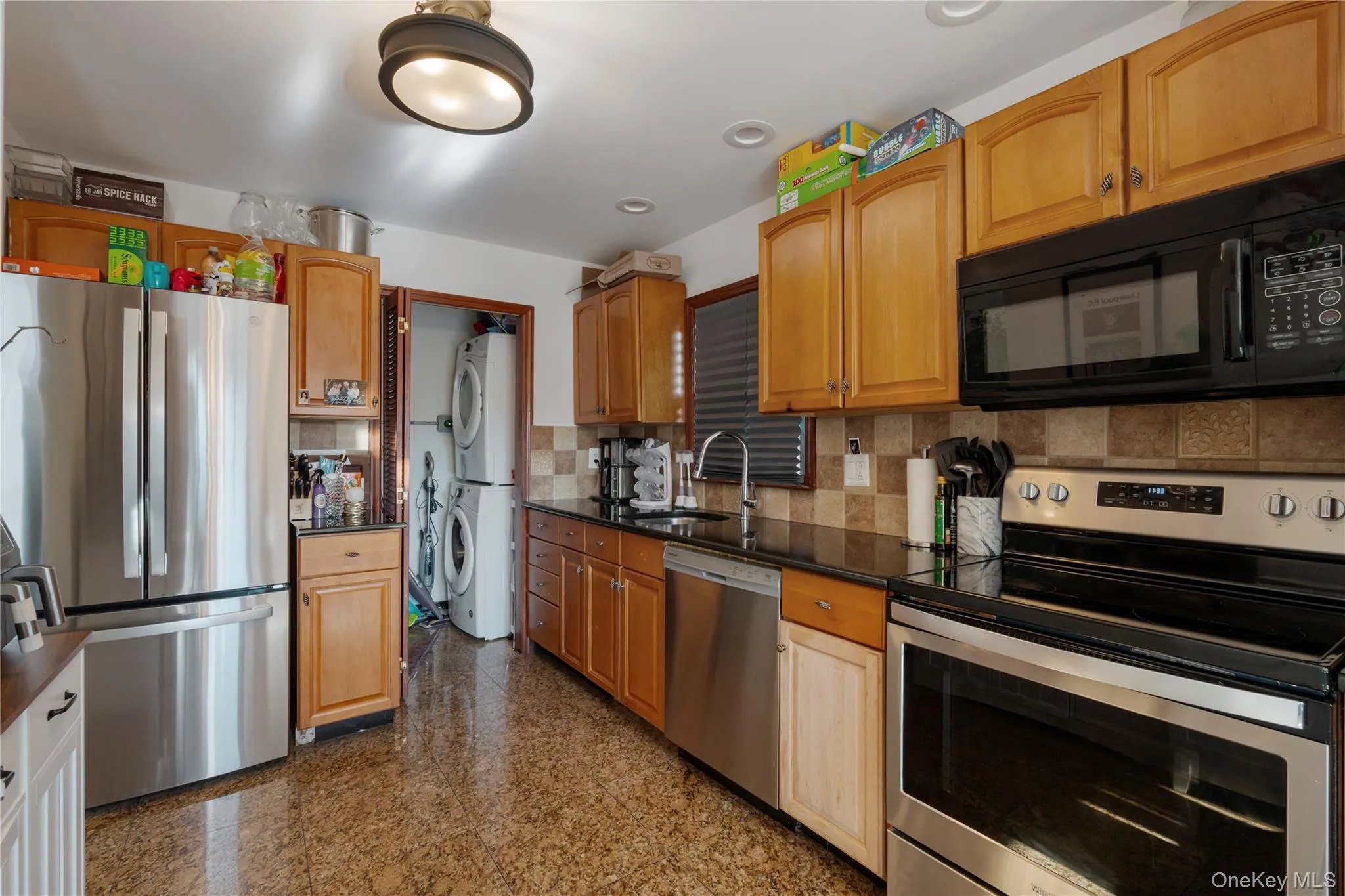 Kitchen with stainless steel appliances, granite tiled floors, stacked washer / dryer, backsplash, and brown cabinetry Kitchen with stainless steel appliances, granite tiled floors, stacked washer / dryer, backsplash, and brown cabinetry