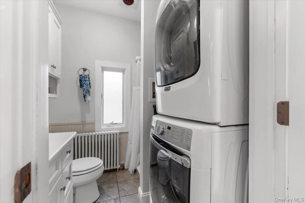 Full bathroom featuring a wainscoted wall, radiator heating unit, curtained shower, stacked washing machine and dryer, and vanity Full bathroom featuring a wainscoted wall, radiator heating unit, curtained shower, stacked washing machine and dryer, and vanity