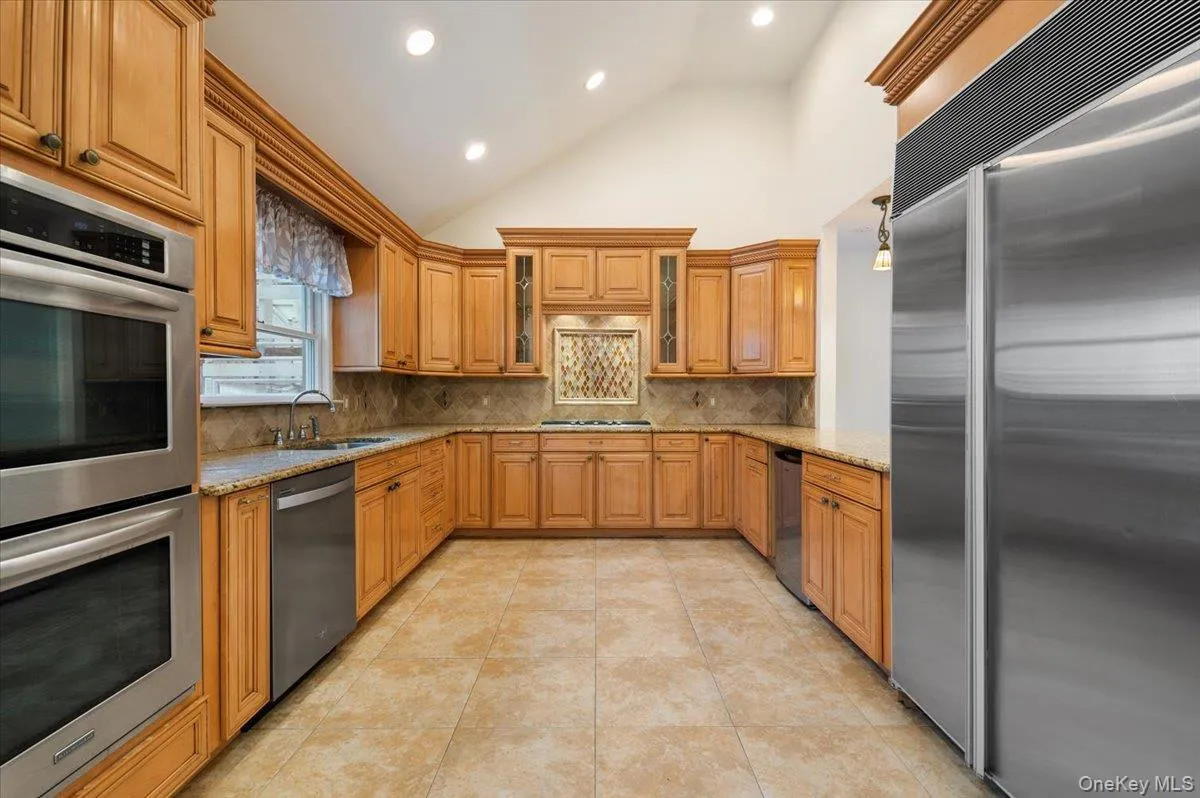 Main Kitchen featuring appliances with stainless steel finishes, light stone countertops, high vaulted ceiling, decorative backsplash, and brown cabinetry Main Kitchen featuring appliances with stainless steel finishes, light stone countertops, high vaulted ceiling, decorative backsplash, and brown cabinetry