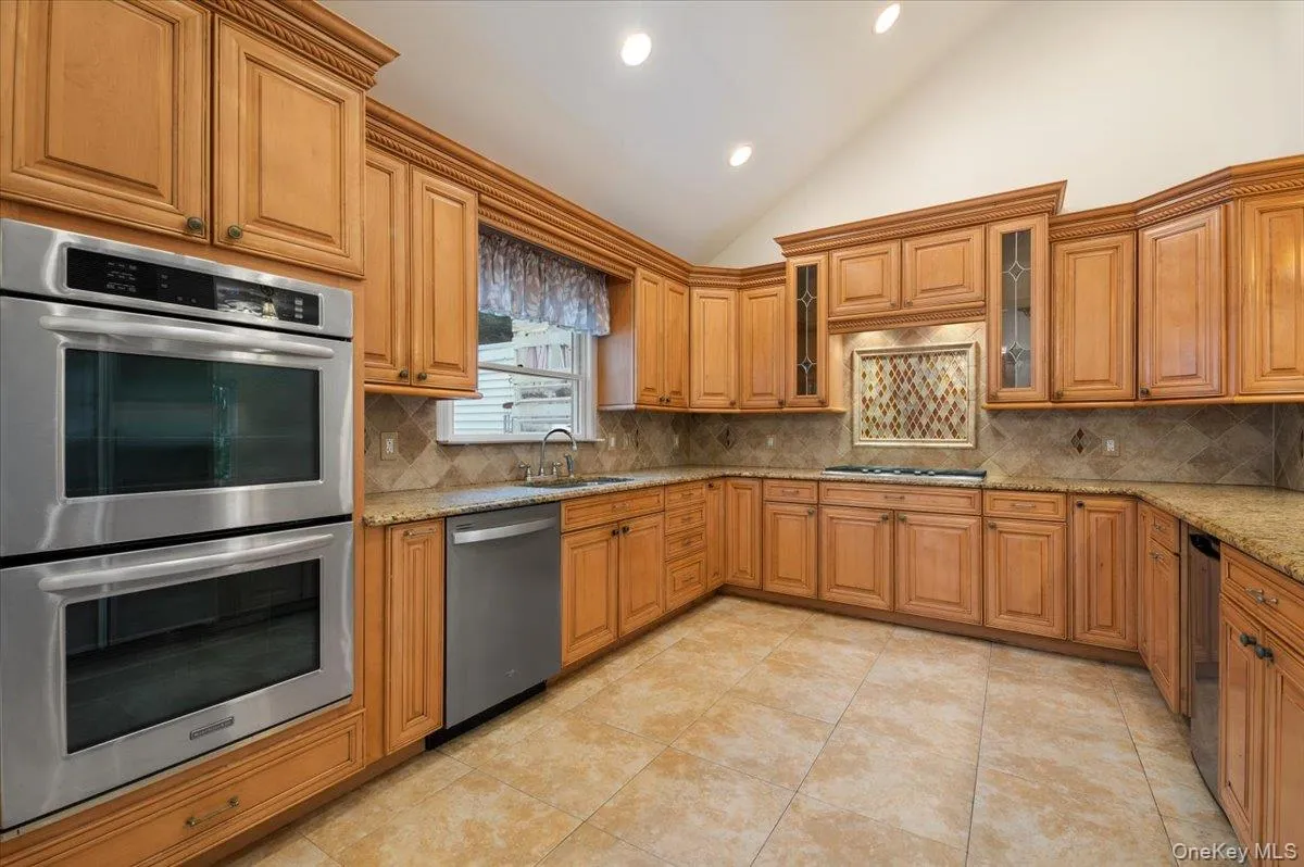 Kitchen with appliances with stainless steel finishes, backsplash, brown cabinetry, high vaulted ceiling, and recessed lighting Kitchen with appliances with stainless steel finishes, backsplash, brown cabinetry, high vaulted ceiling, and recessed lighting