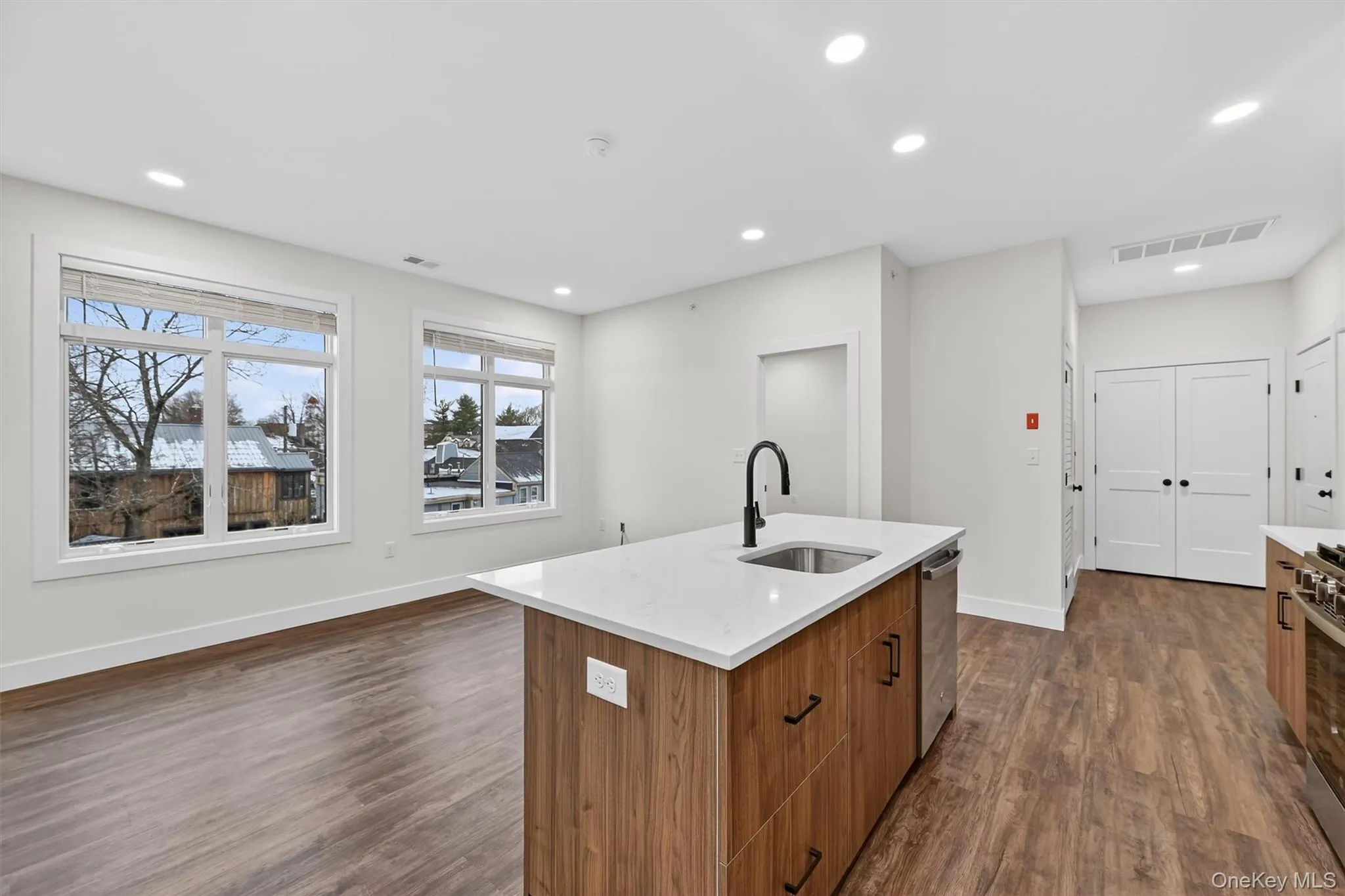 Kitchen featuring brown cabinetry, dark wood-style floors, light stone counters, an island with sink, and recessed lighting Kitchen featuring brown cabinetry, dark wood-style floors, light stone counters, an island with sink, and recessed lighting