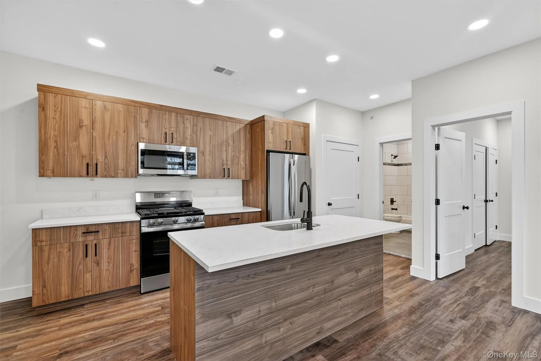 Kitchen featuring appliances with stainless steel finishes, brown cabinetry, a center island with sink, dark wood finished floors, and recessed lighting Kitchen featuring appliances with stainless steel finishes, brown cabinetry, a center island with sink, dark wood finished floors, and recessed lighting