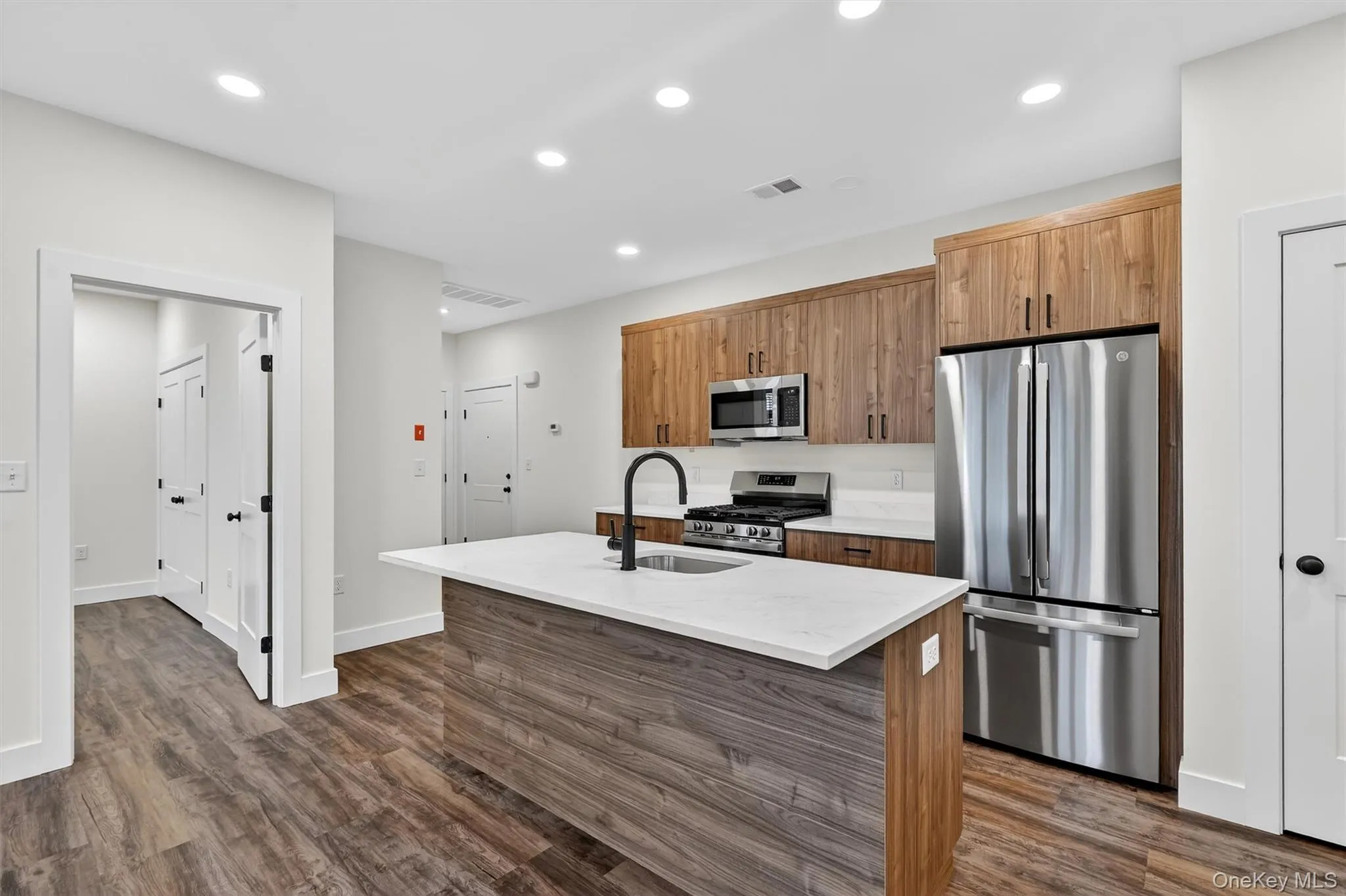 Kitchen with stainless steel appliances, brown cabinetry, an island with sink, dark wood-style floors, and recessed lighting Kitchen with stainless steel appliances, brown cabinetry, an island with sink, dark wood-style floors, and recessed lighting