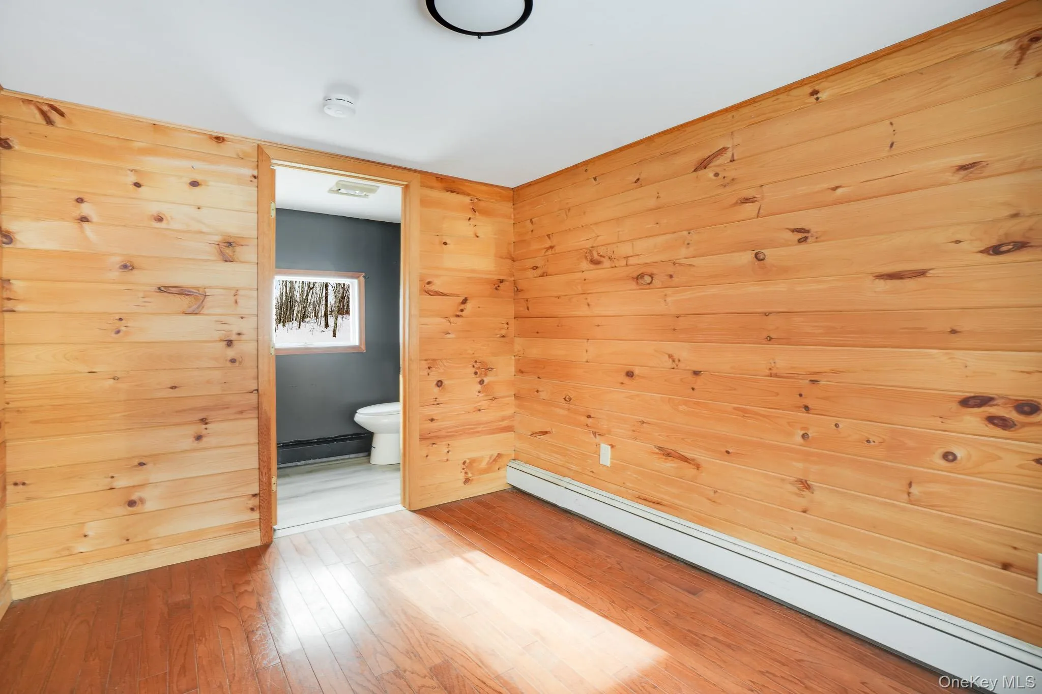 Empty room with baseboard heating, wood walls, light wood-style floors, and a baseboard radiator Empty room with baseboard heating, wood walls, light wood-style floors, and a baseboard radiator