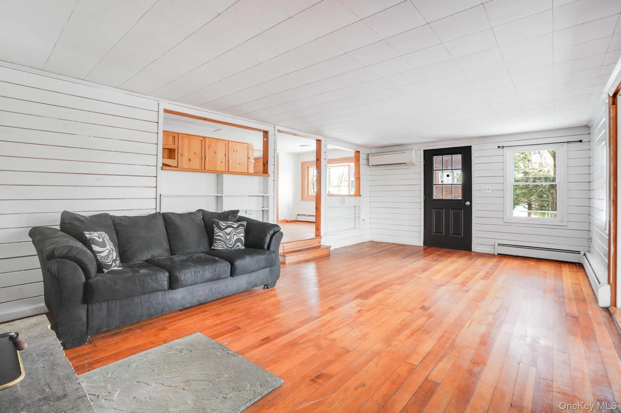 Living area featuring wooden walls, light wood-type flooring, plenty of natural light, a wall mounted air conditioner, and baseboard heating Living area featuring wooden walls, light wood-type flooring, plenty of natural light, a wall mounted air conditioner, and baseboard heating