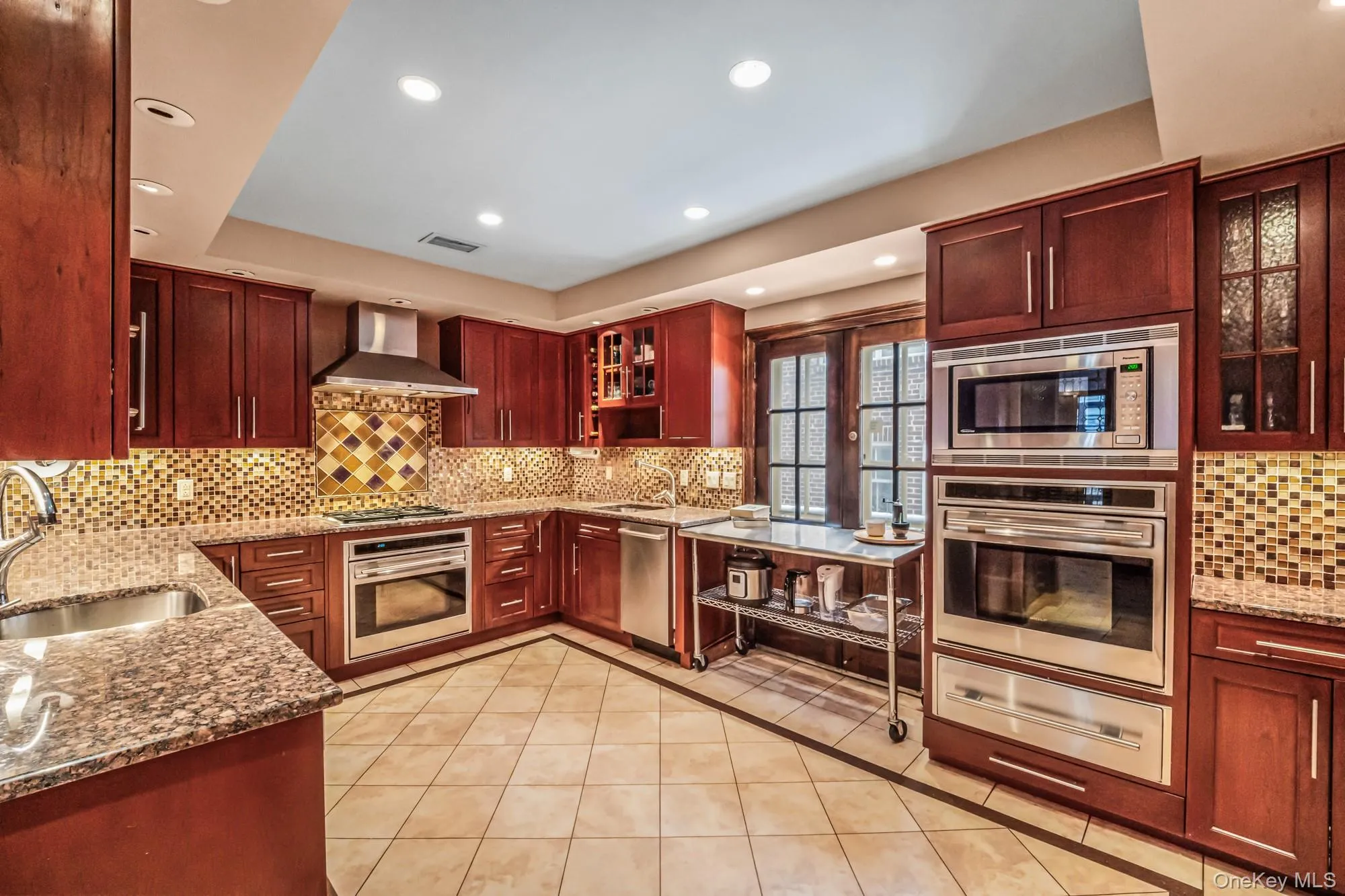 Kitchen featuring reddish brown cabinets, dark stone countertops, glass insert cabinets, appliances with stainless steel finishes, and wall chimney range hood Kitchen featuring reddish brown cabinets, dark stone countertops, glass insert cabinets, appliances with stainless steel finishes, and wall chimney range hood
