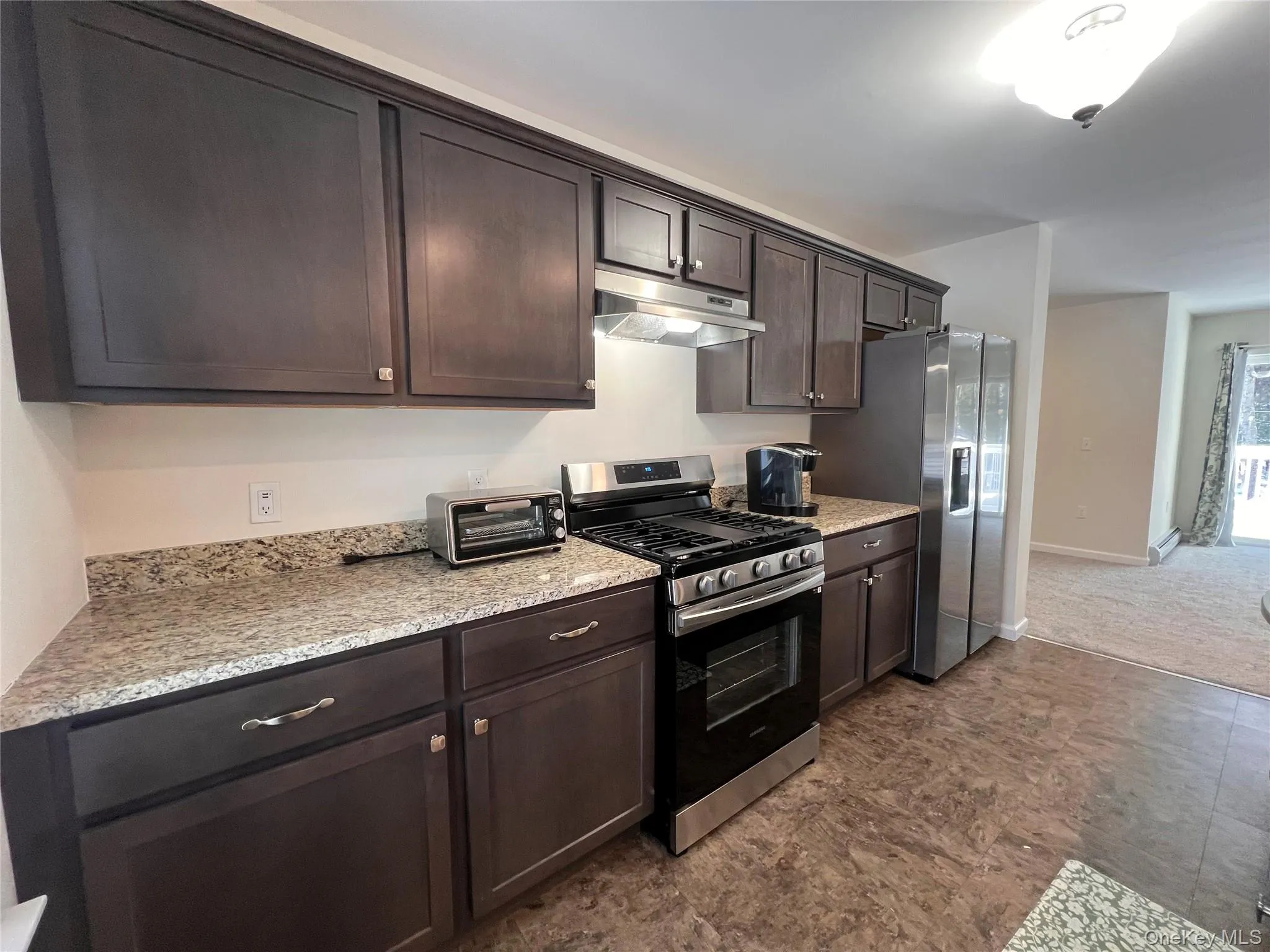 Kitchen featuring stainless steel appliances, dark brown cabinets, under cabinet range hood, and light stone countertops Kitchen featuring stainless steel appliances, dark brown cabinets, under cabinet range hood, and light stone countertops
