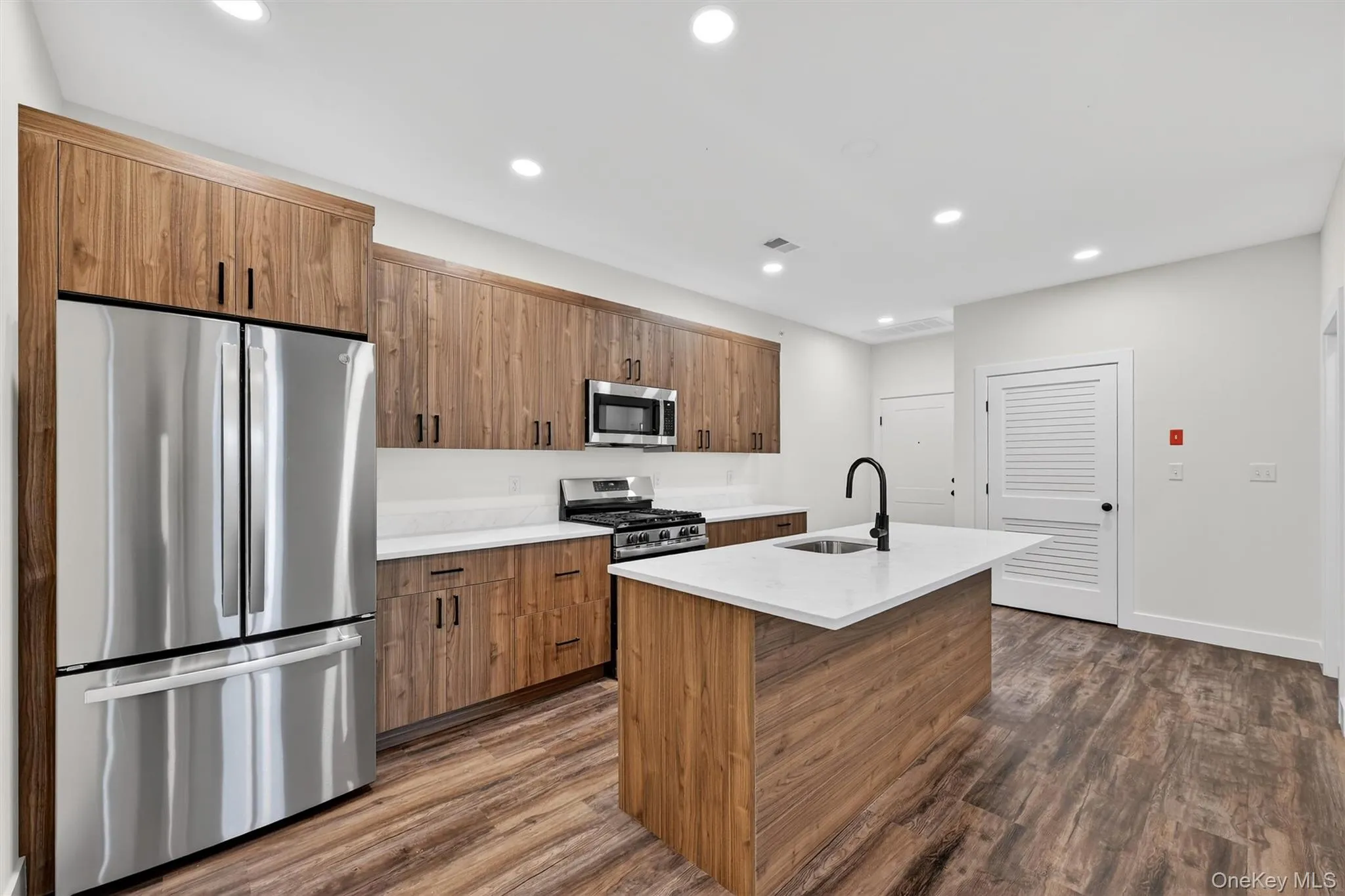 Kitchen featuring stainless steel appliances, a center island with sink, recessed lighting, brown cabinetry, and dark wood-style floors Kitchen featuring stainless steel appliances, a center island with sink, recessed lighting, brown cabinetry, and dark wood-style floors
