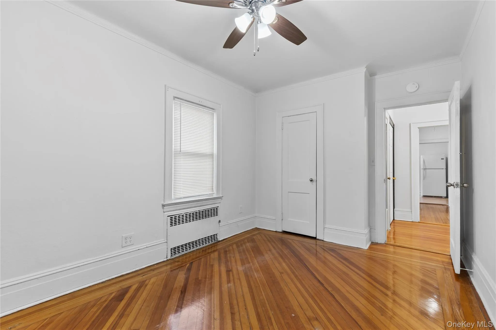 Unfurnished bedroom with radiator, freestanding refrigerator, a ceiling fan, wood-type flooring, and crown molding Unfurnished bedroom with radiator, freestanding refrigerator, a ceiling fan, wood-type flooring, and crown molding