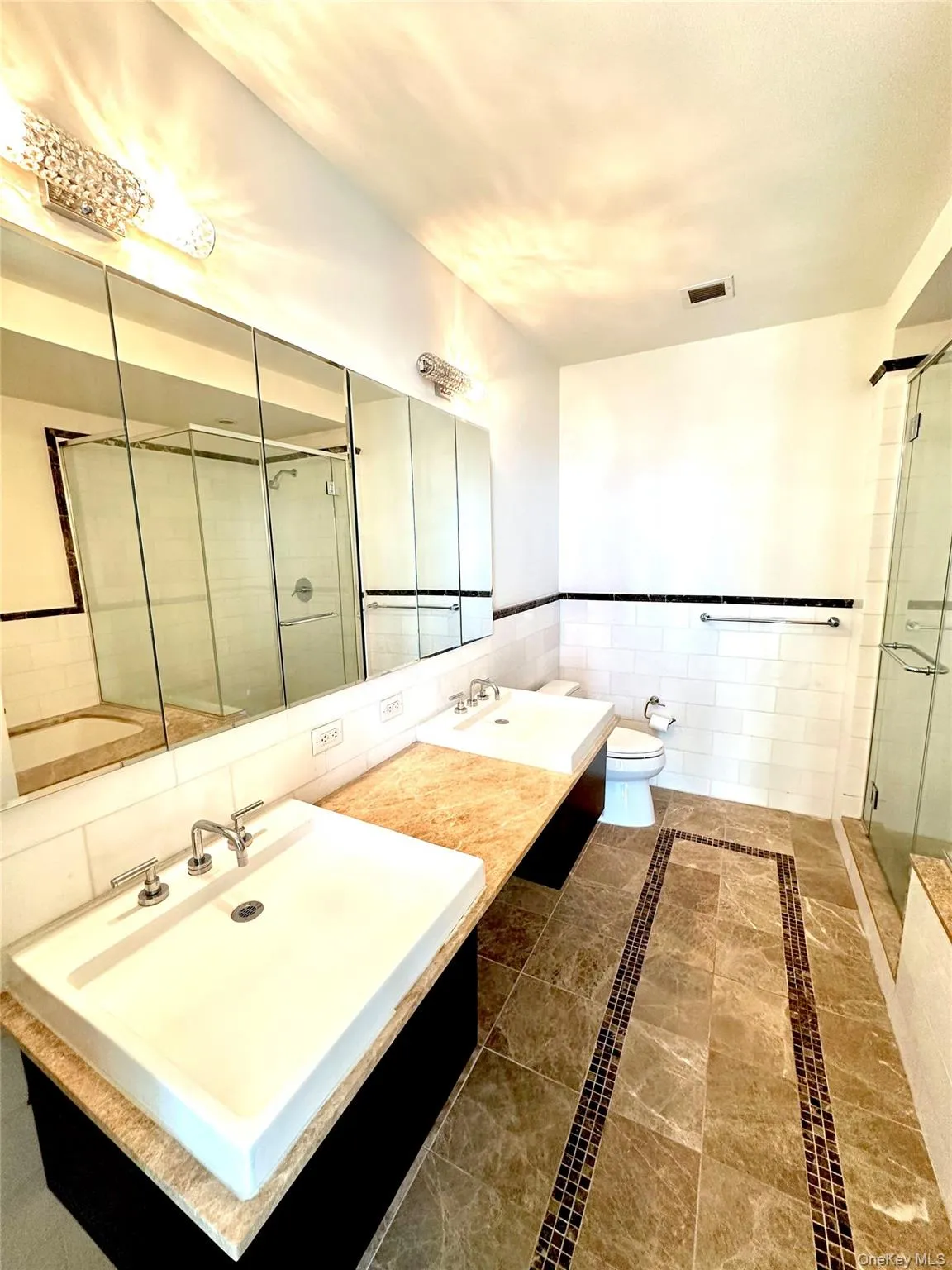 Full bath with double vanity, inlaid floor details, a stall shower, tile walls, and a wainscoted wall Full bath with double vanity, inlaid floor details, a stall shower, tile walls, and a wainscoted wall