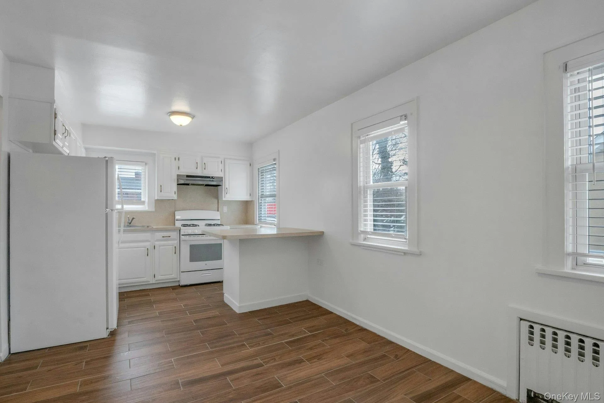 Kitchen with radiator heating unit, white appliances, light countertops, white cabinetry, and wood tiled floors Kitchen with radiator heating unit, white appliances, light countertops, white cabinetry, and wood tiled floors