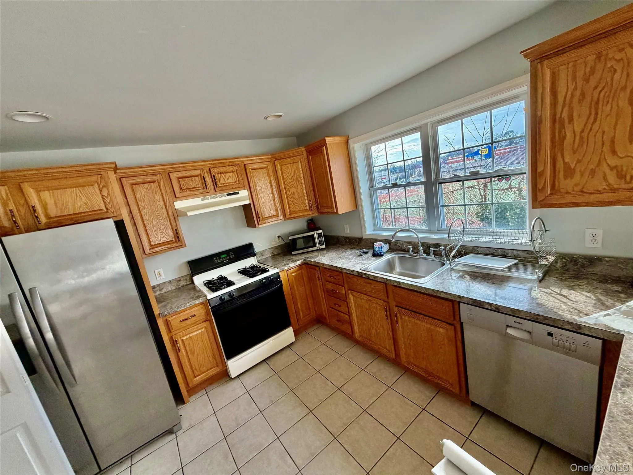 Kitchen featuring stainless steel appliances, under cabinet range hood, light stone countertops, brown cabinets, and light tile patterned floors Kitchen featuring stainless steel appliances, under cabinet range hood, light stone countertops, brown cabinets, and light tile patterned floors