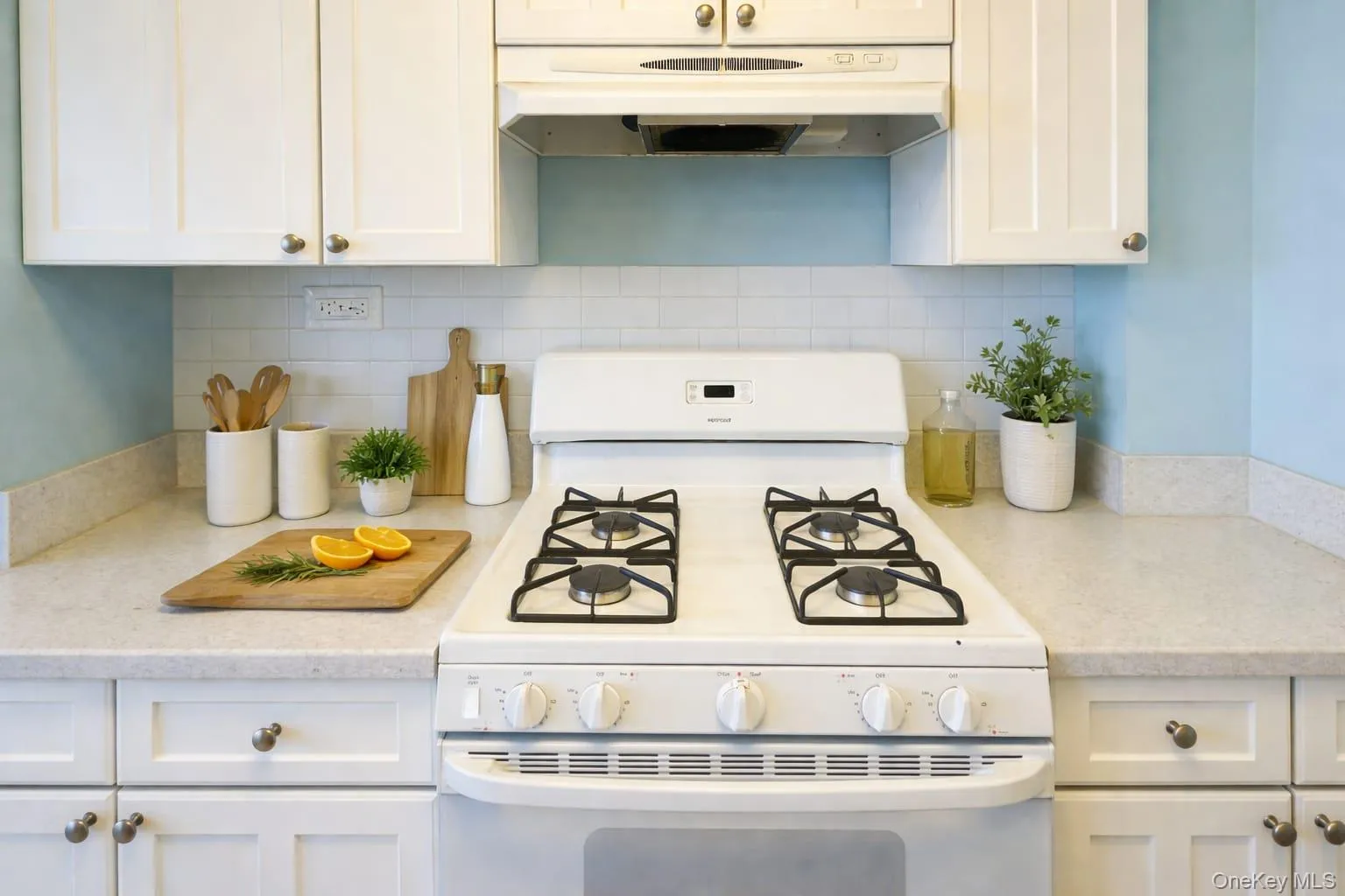 Kitchen with white gas range oven, white cabinetry, under cabinet range hood, light stone counters, and backsplash Kitchen with white gas range oven, white cabinetry, under cabinet range hood, light stone counters, and backsplash