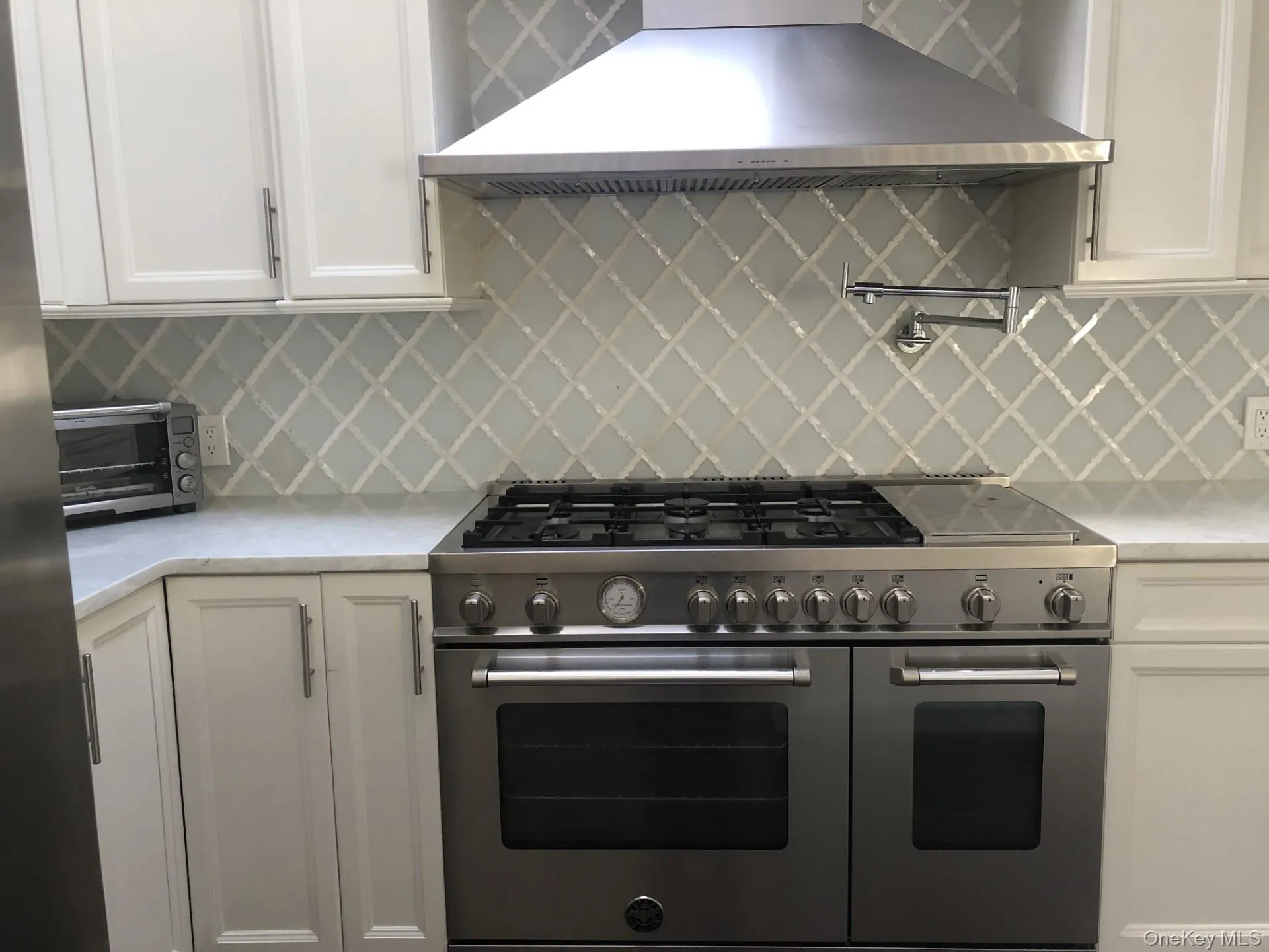 Kitchen with a toaster, white cabinetry, decorative backsplash, range hood, and range with two ovens Kitchen with a toaster, white cabinetry, decorative backsplash, range hood, and range with two ovens