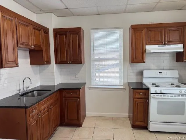 Kitchen featuring white range with electric cooktop, under cabinet range hood, backsplash, brown cabinets, and a paneled ceiling Kitchen featuring white range with electric cooktop, under cabinet range hood, backsplash, brown cabinets, and a paneled ceiling