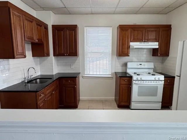 Kitchen with white appliances, under cabinet range hood, backsplash, light tile patterned flooring, and a paneled ceiling Kitchen with white appliances, under cabinet range hood, backsplash, light tile patterned flooring, and a paneled ceiling