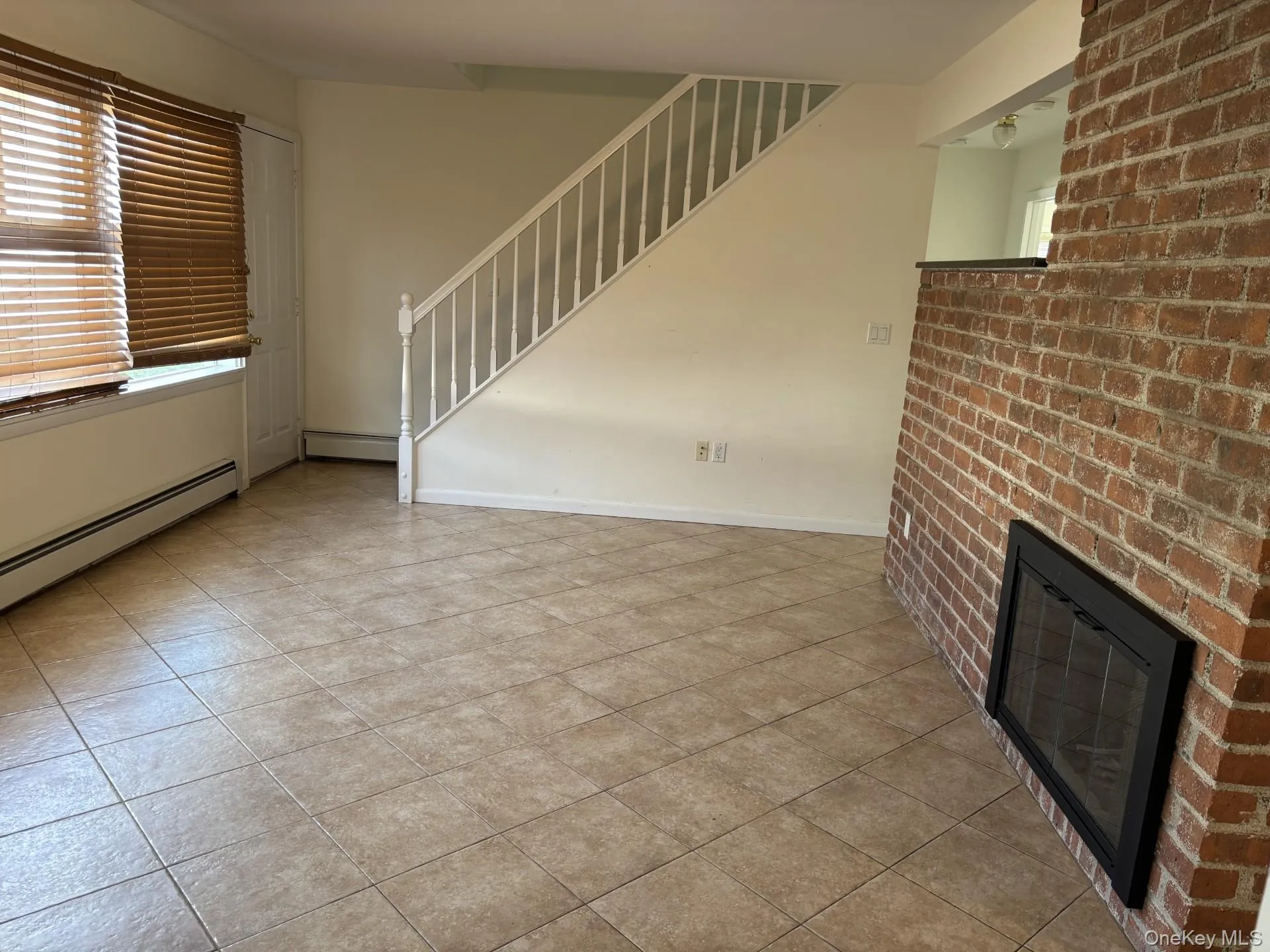 Unfurnished living room featuring baseboard heating, a fireplace, stairway, light tile patterned floors, and brick wall Unfurnished living room featuring baseboard heating, a fireplace, stairway, light tile patterned floors, and brick wall