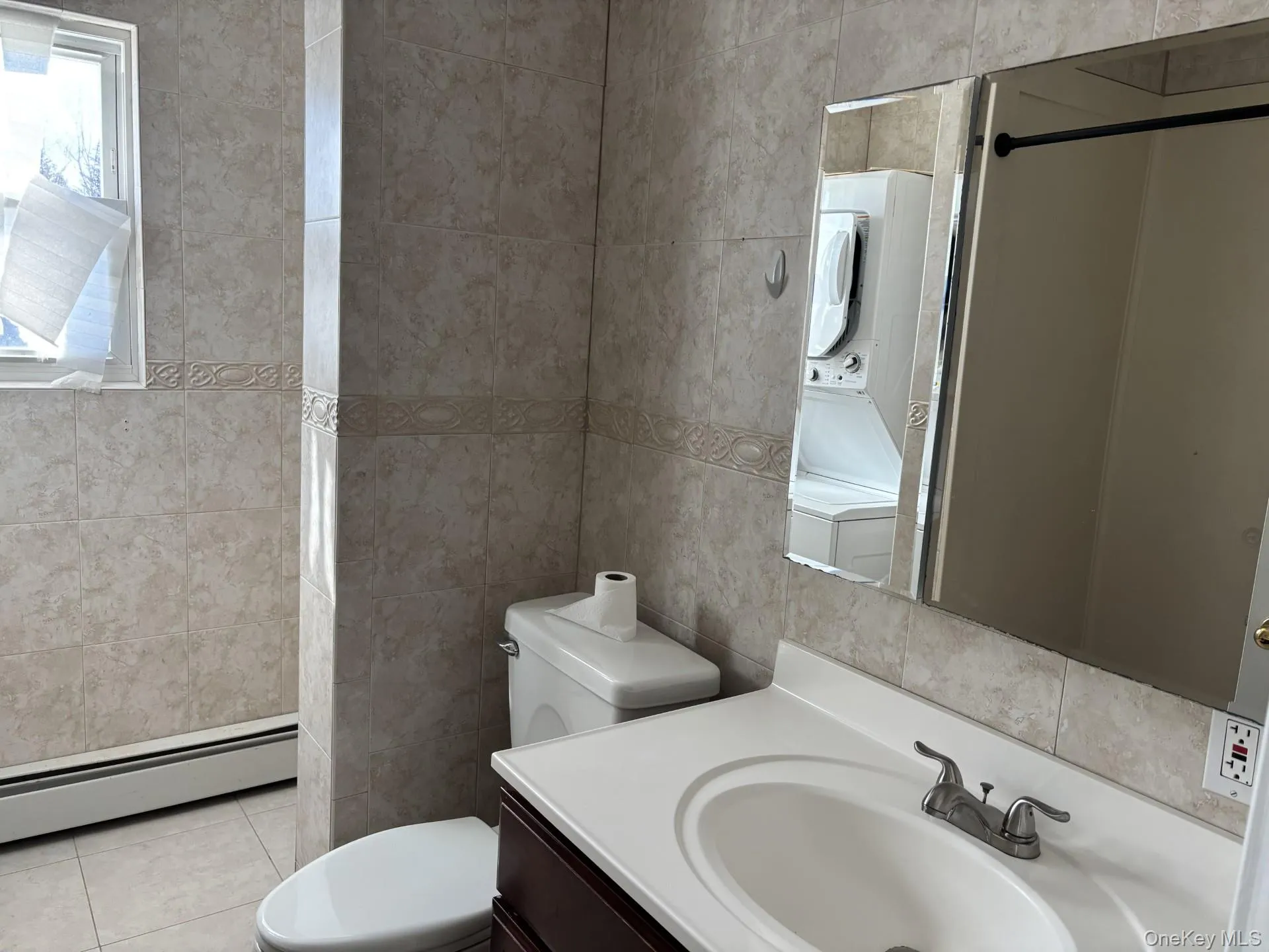 Bathroom with vanity, a baseboard radiator, tile walls, light tile patterned floors, and stacked washer and clothes dryer Bathroom with vanity, a baseboard radiator, tile walls, light tile patterned floors, and stacked washer and clothes dryer
