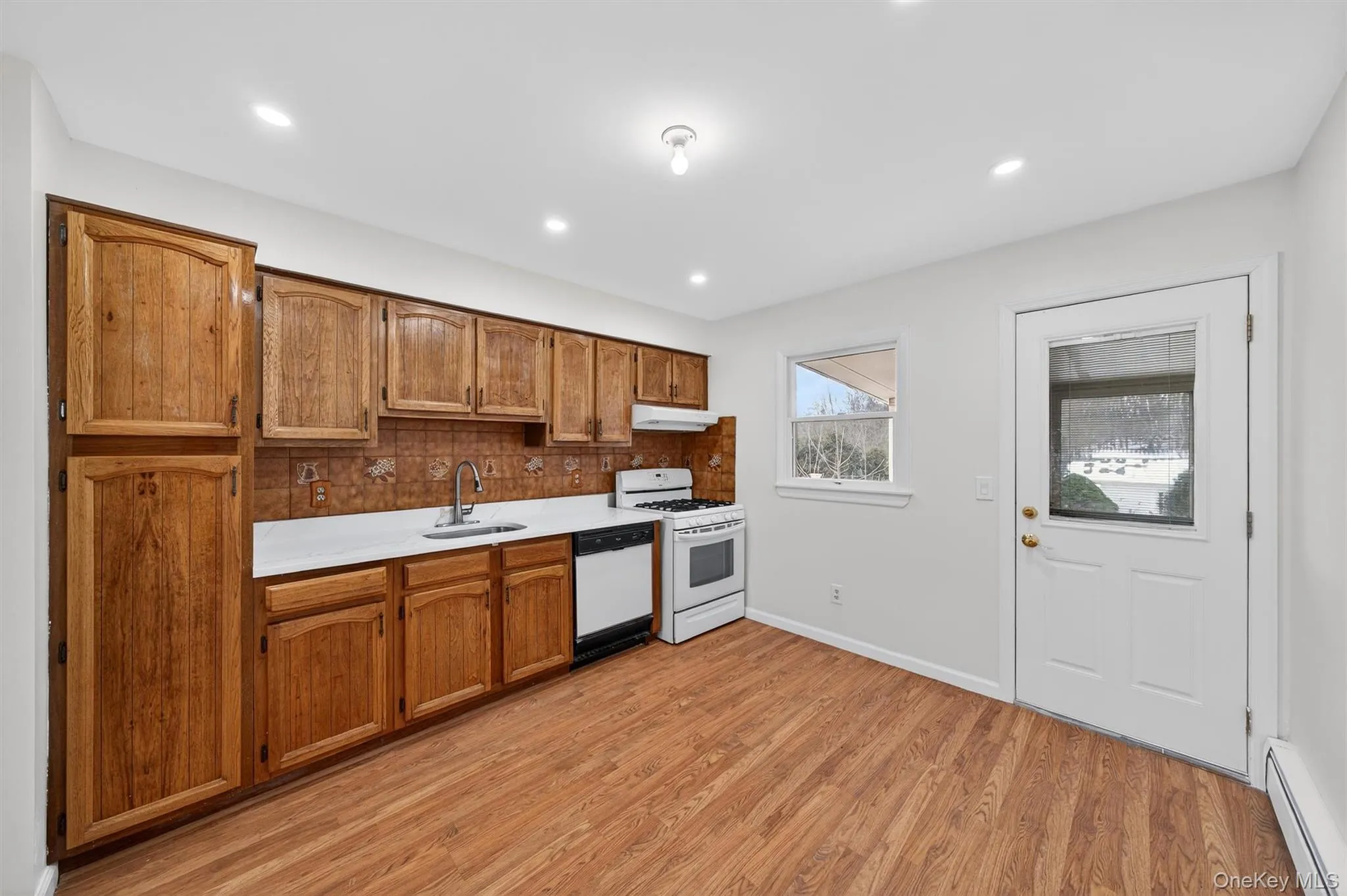 Kitchen with light countertops, white appliances, baseboard heating, brown cabinets, and recessed lighting Kitchen with light countertops, white appliances, baseboard heating, brown cabinets, and recessed lighting