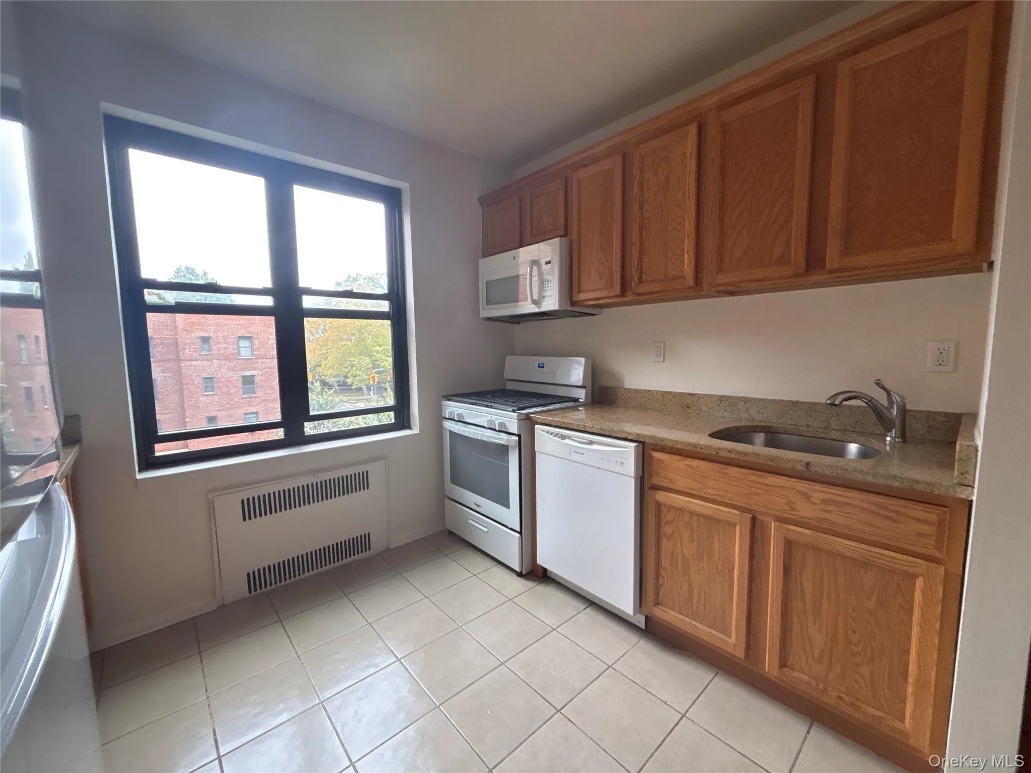 Kitchen featuring white appliances, radiator heating unit, light stone counters, brown cabinets, and light tile patterned flooring Kitchen featuring white appliances, radiator heating unit, light stone counters, brown cabinets, and light tile patterned flooring