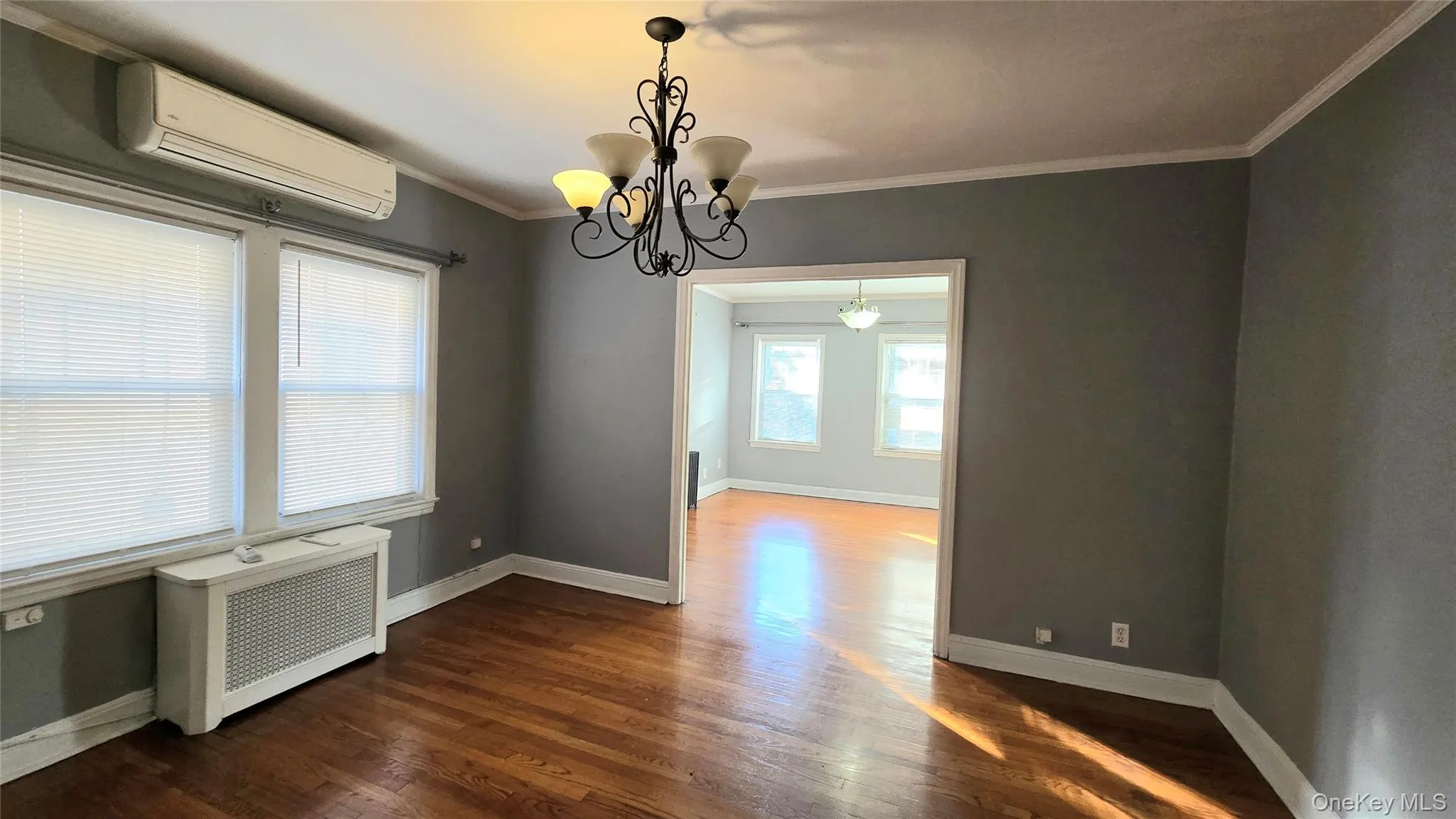 Unfurnished dining area with radiator heating unit, a wall mounted AC, ornamental molding, a chandelier, and dark wood-style floors Unfurnished dining area with radiator heating unit, a wall mounted AC, ornamental molding, a chandelier, and dark wood-style floors