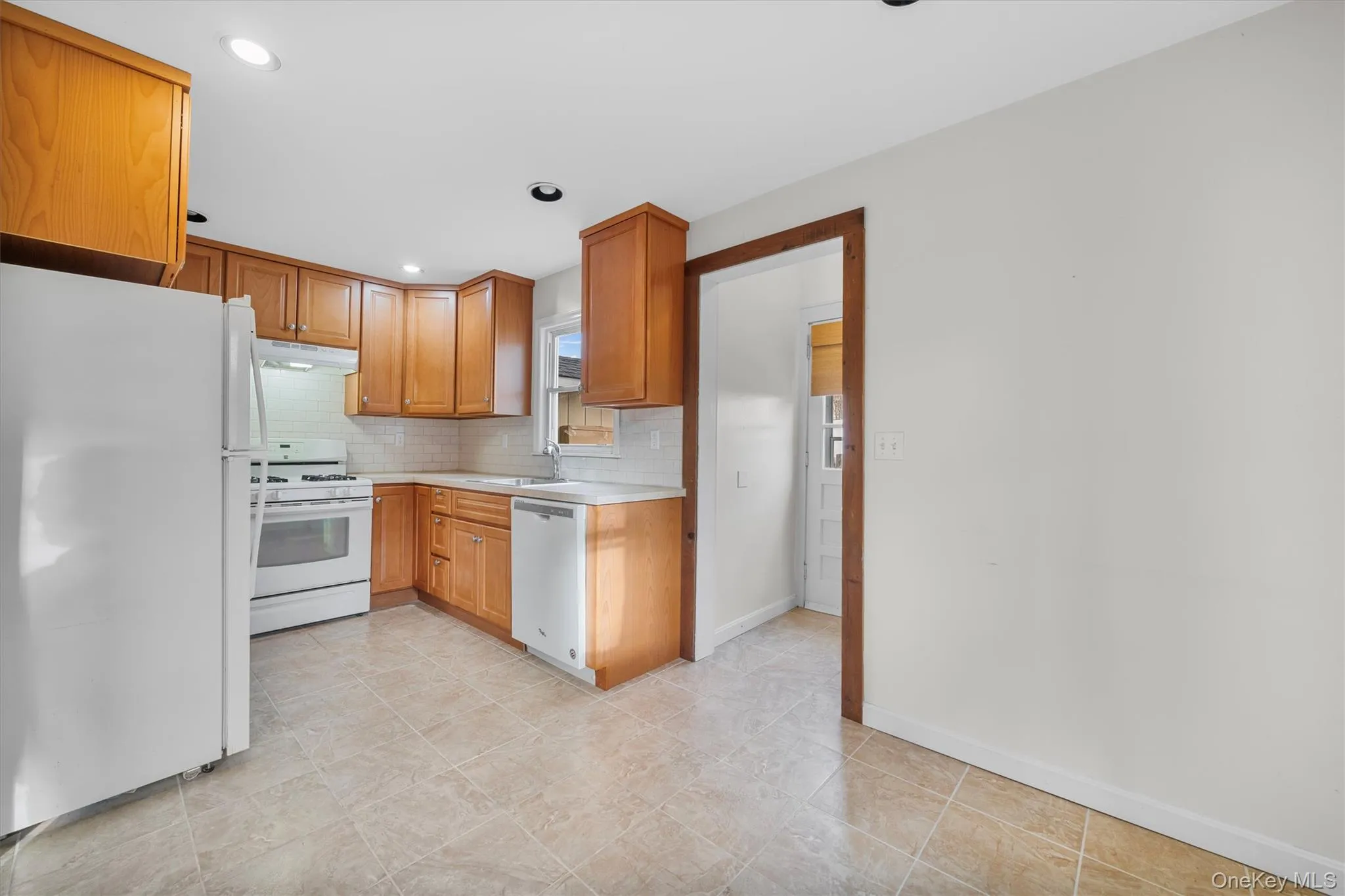 Kitchen with white appliances, light countertops, backsplash, brown cabinets, and under cabinet range hood Kitchen with white appliances, light countertops, backsplash, brown cabinets, and under cabinet range hood