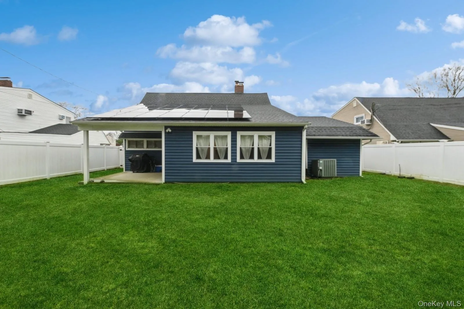 Rear view of house with roof mounted solar panels, a patio area, roof with shingles, a fenced backyard, and a chimney Rear view of house with roof mounted solar panels, a patio area, roof with shingles, a fenced backyard, and a chimney