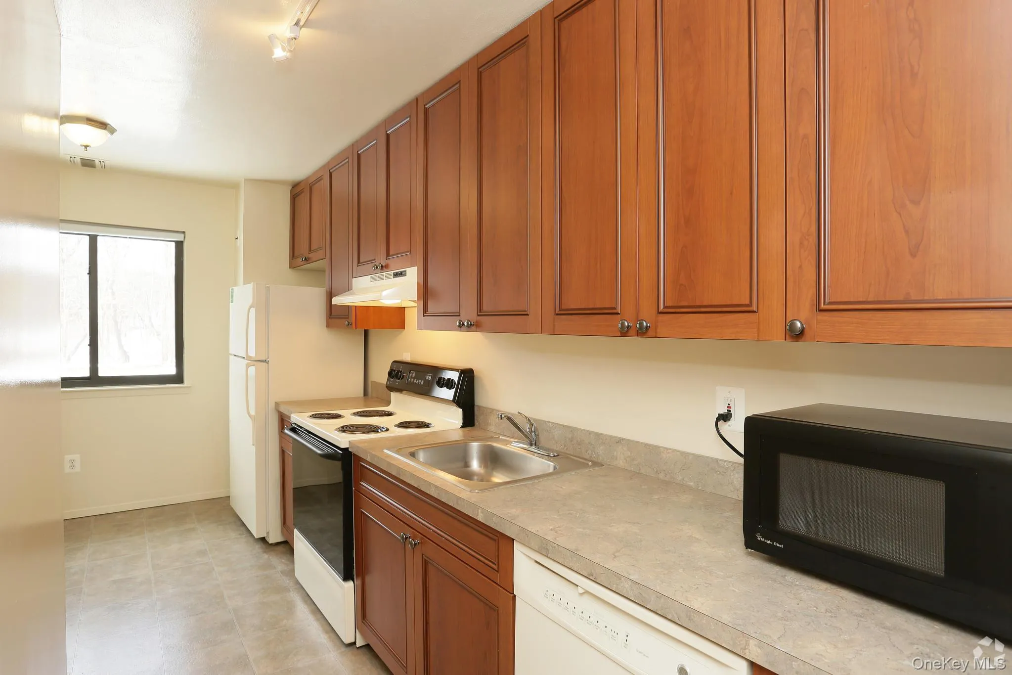 Kitchen featuring white appliances, brown cabinets, light countertops, under cabinet range hood, and track lighting Kitchen featuring white appliances, brown cabinets, light countertops, under cabinet range hood, and track lighting