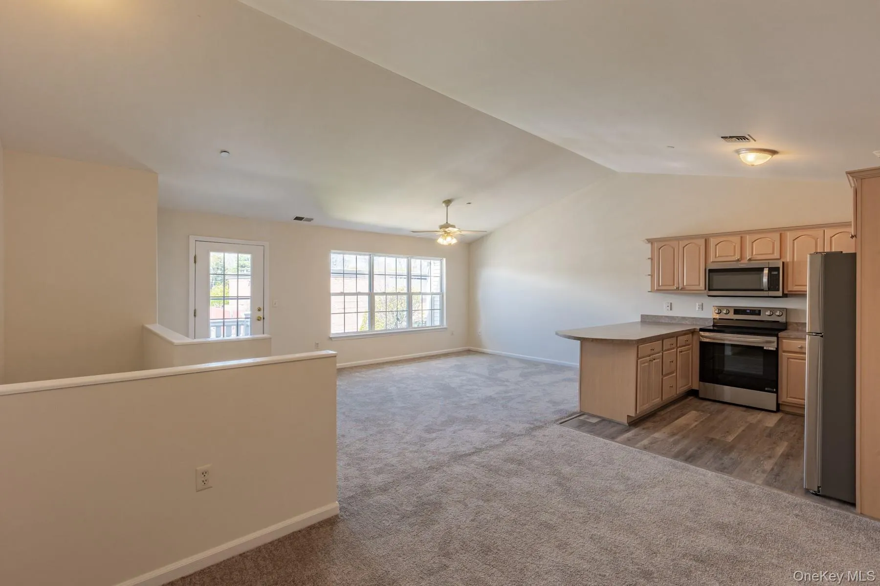 Kitchen with light brown cabinetry, lofted ceiling, appliances with stainless steel finishes, open floor plan, and dark carpet Kitchen with light brown cabinetry, lofted ceiling, appliances with stainless steel finishes, open floor plan, and dark carpet
