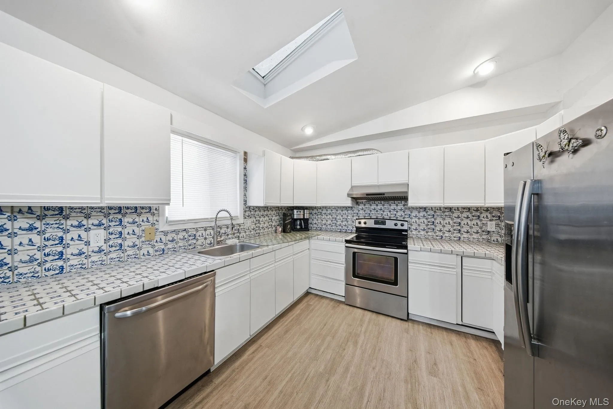 Kitchen featuring tile counters, vaulted ceiling, appliances with stainless steel finishes, white cabinets, and a skylight Kitchen featuring tile counters, vaulted ceiling, appliances with stainless steel finishes, white cabinets, and a skylight