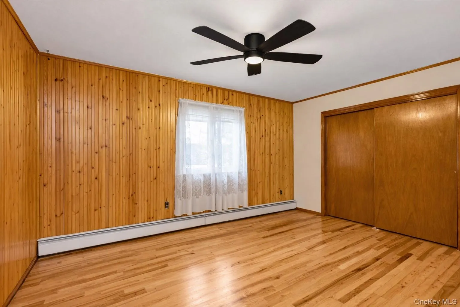 Unfurnished bedroom featuring a baseboard heating unit, light wood-style floors, wood walls, crown molding, and a closet Unfurnished bedroom featuring a baseboard heating unit, light wood-style floors, wood walls, crown molding, and a closet