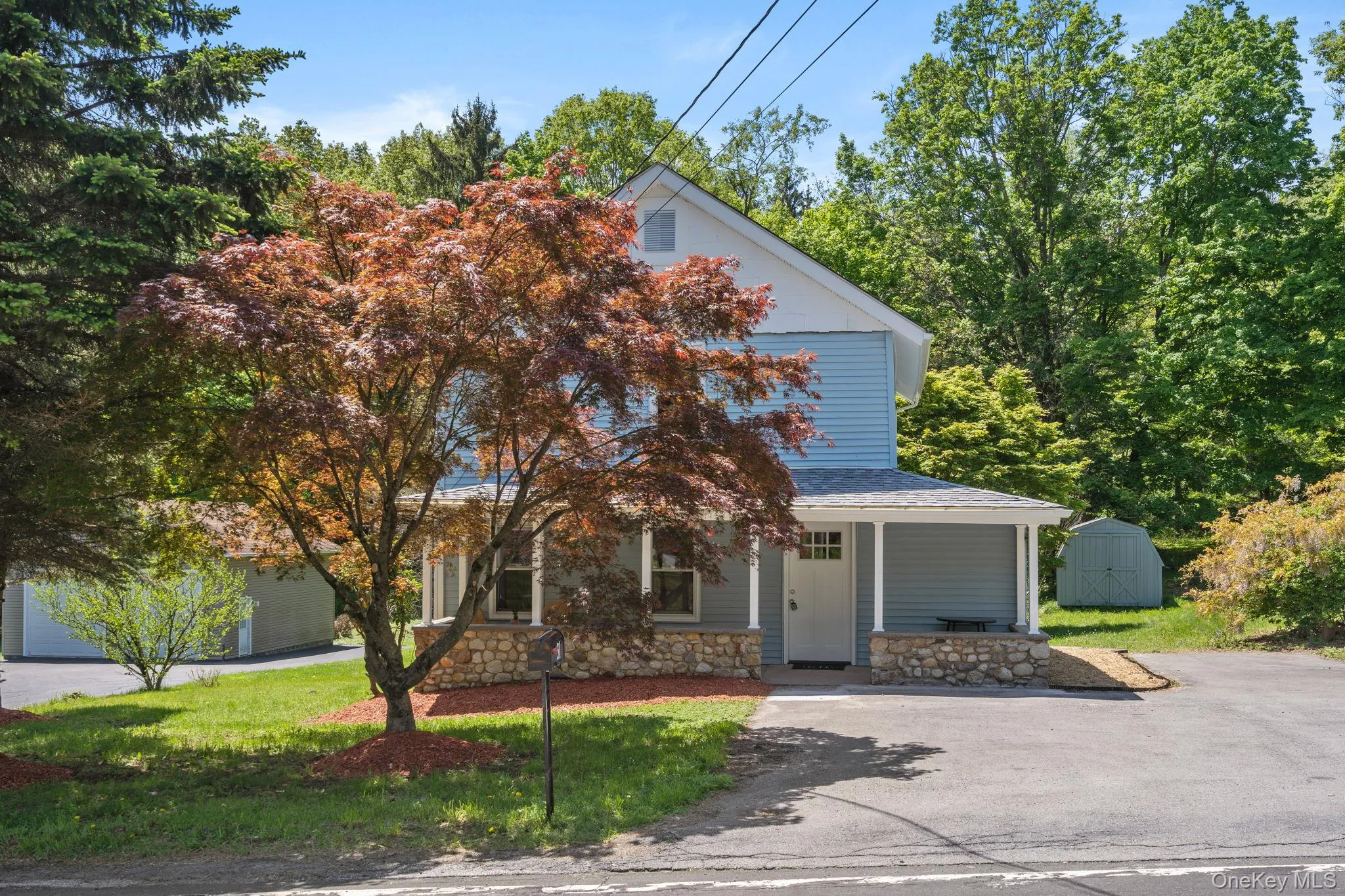 View of front of home featuring stone siding, a front lawn, covered porch, a shingled roof, and a storage unit View of front of home featuring stone siding, a front lawn, covered porch, a shingled roof, and a storage unit