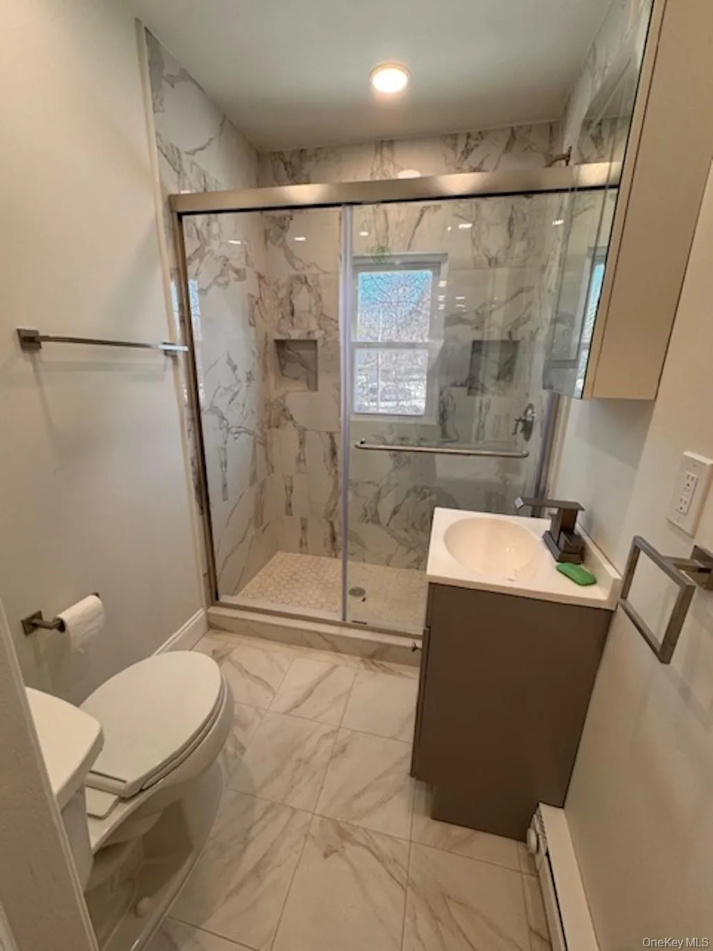 Full bathroom with light marble finish flooring, vanity, a marble finish shower, and a baseboard heating unit Full bathroom with light marble finish flooring, vanity, a marble finish shower, and a baseboard heating unit