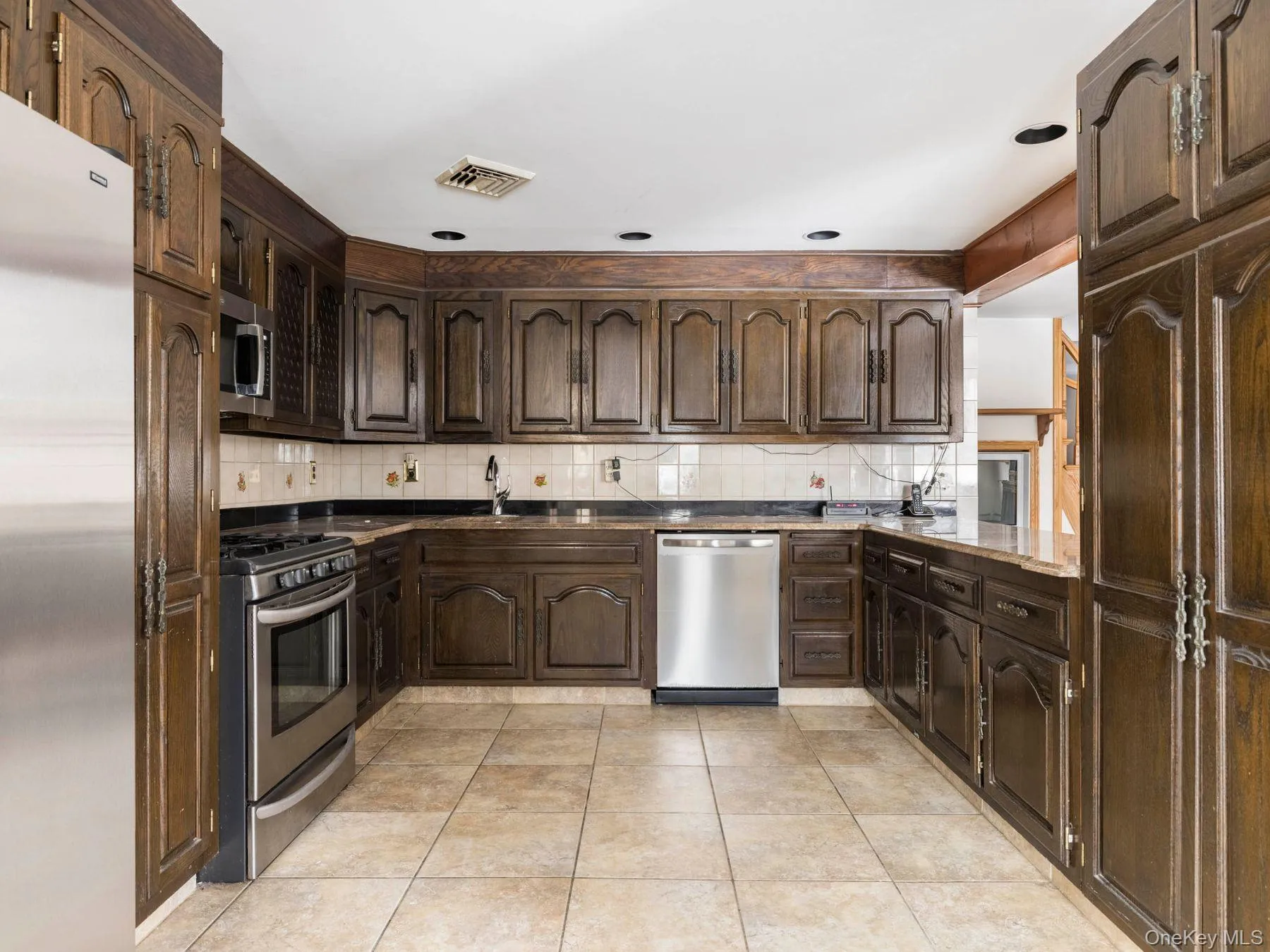 Kitchen featuring dark brown cabinets, stainless steel appliances, tasteful backsplash, light tile patterned flooring, and dark stone counters Kitchen featuring dark brown cabinets, stainless steel appliances, tasteful backsplash, light tile patterned flooring, and dark stone counters