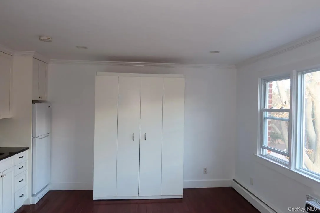 Unfurnished bedroom with crown molding, freestanding refrigerator, dark wood-style flooring, baseboard heating, and a closet Unfurnished bedroom with crown molding, freestanding refrigerator, dark wood-style flooring, baseboard heating, and a closet