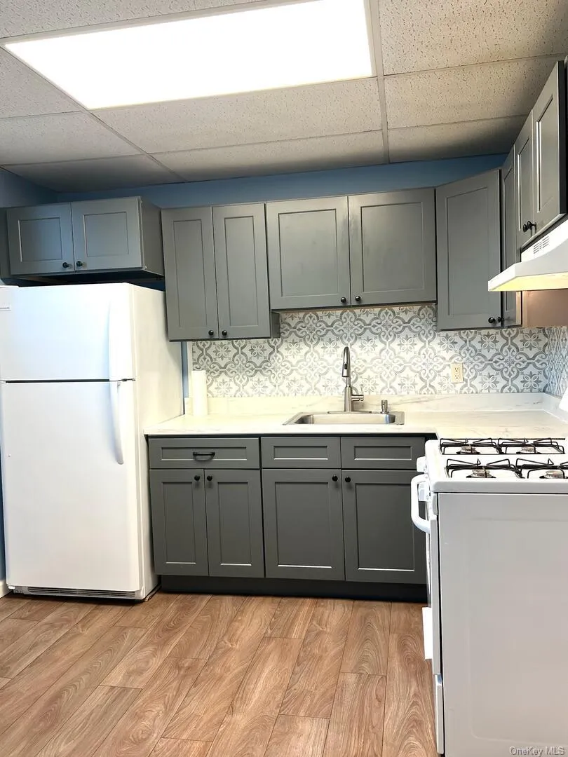 Kitchen featuring a paneled ceiling, white appliances, light wood-type flooring, gray cabinets, and under cabinet range hood Kitchen featuring a paneled ceiling, white appliances, light wood-type flooring, gray cabinets, and under cabinet range hood
