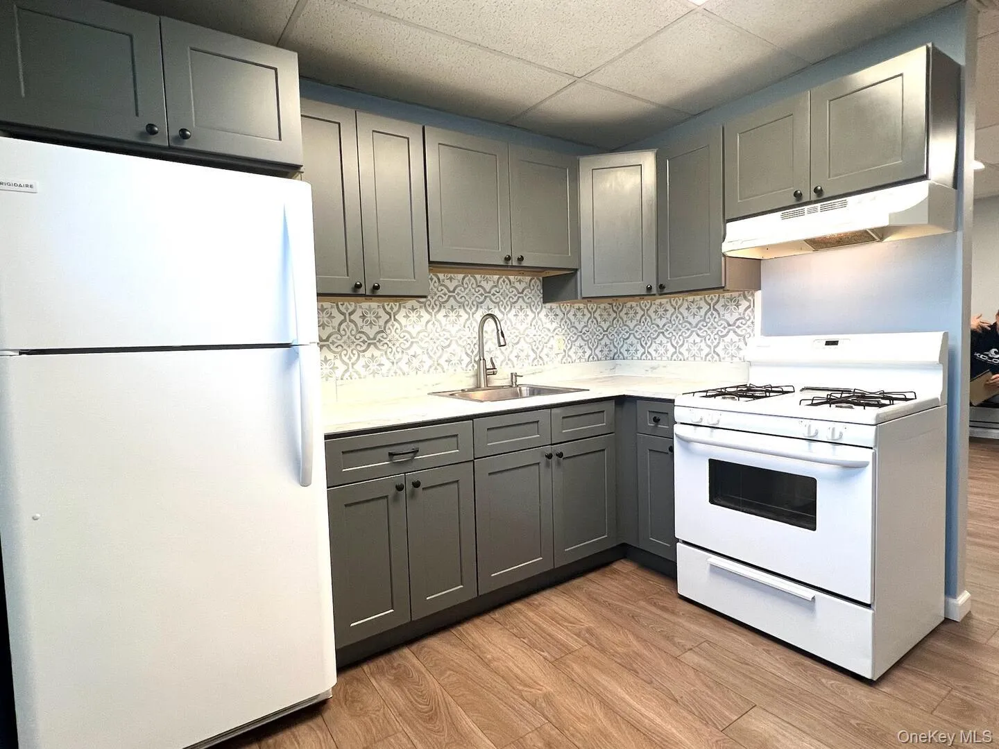 Kitchen featuring white appliances, a drop ceiling, gray cabinets, light countertops, and under cabinet range hood Kitchen featuring white appliances, a drop ceiling, gray cabinets, light countertops, and under cabinet range hood
