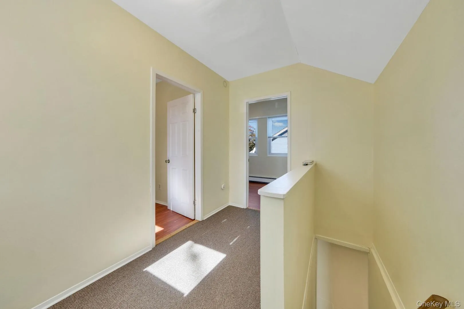 Corridor with an upstairs landing, dark colored carpet, lofted ceiling, and a baseboard radiator Corridor with an upstairs landing, dark colored carpet, lofted ceiling, and a baseboard radiator