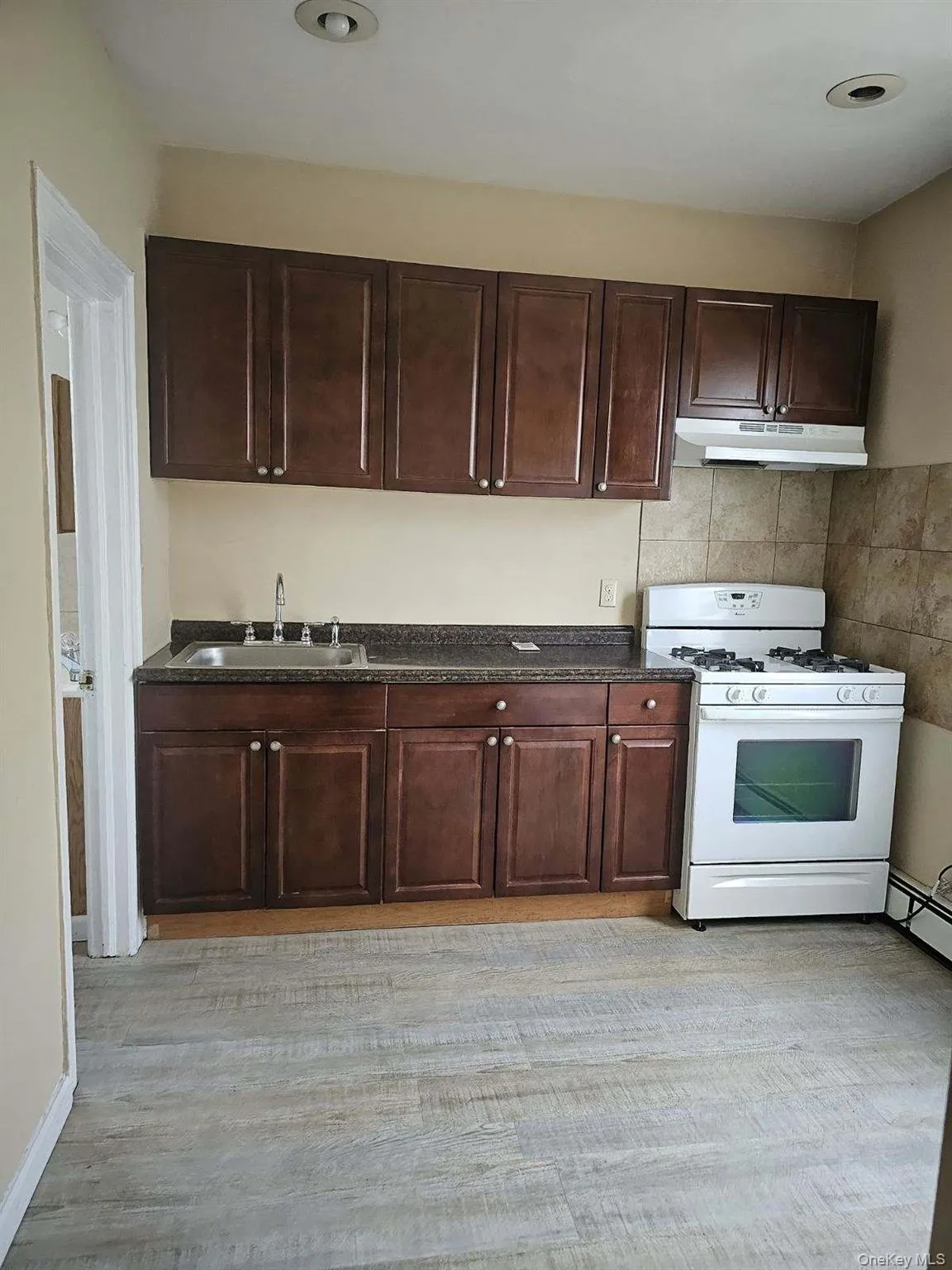 Kitchen featuring dark brown cabinets, white gas stove, dark countertops, under cabinet range hood, and a baseboard radiator Kitchen featuring dark brown cabinets, white gas stove, dark countertops, under cabinet range hood, and a baseboard radiator