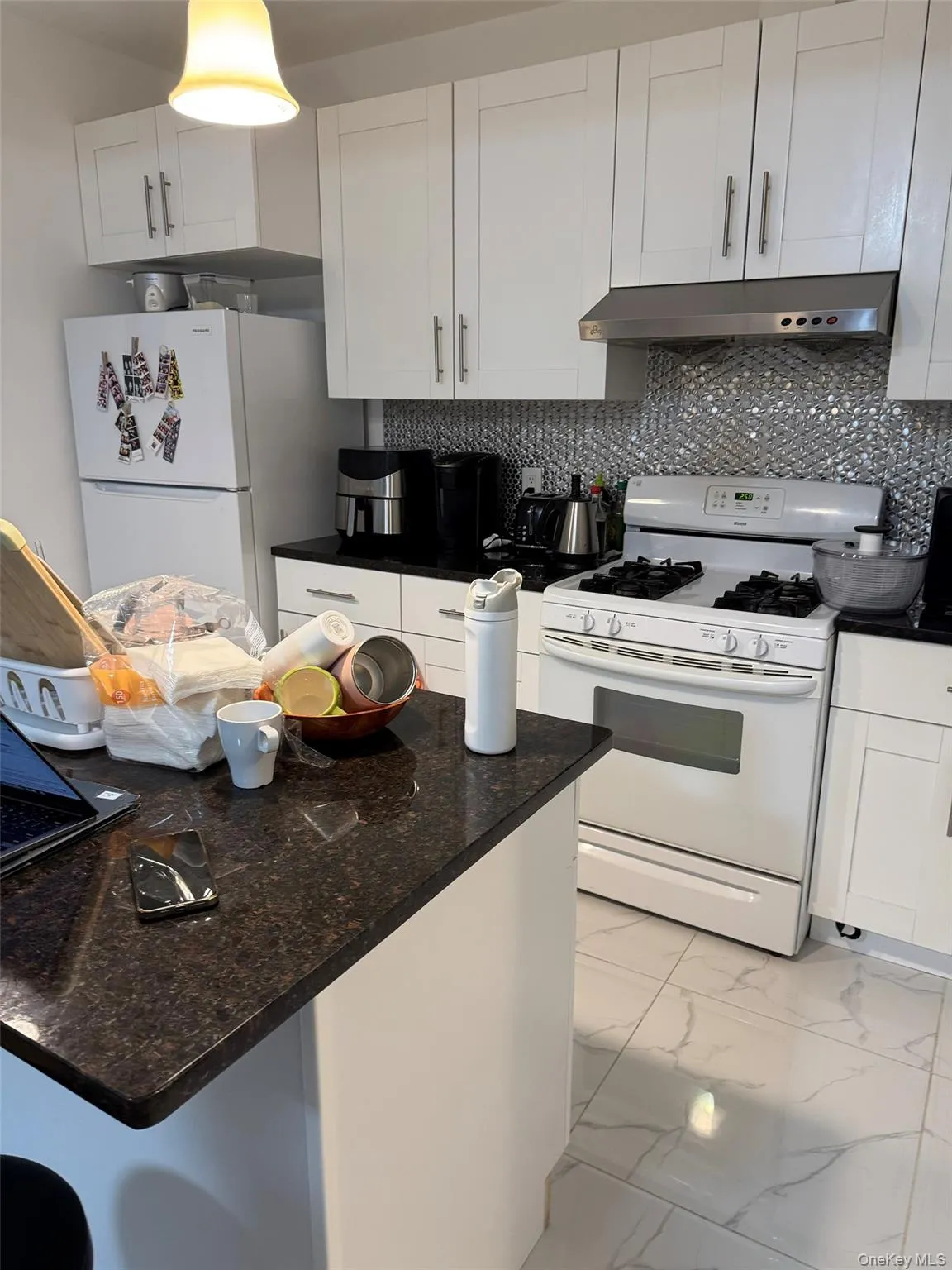 Kitchen featuring white appliances, wall chimney range hood, light marble finish floors, decorative backsplash, and white cabinetry Kitchen featuring white appliances, wall chimney range hood, light marble finish floors, decorative backsplash, and white cabinetry