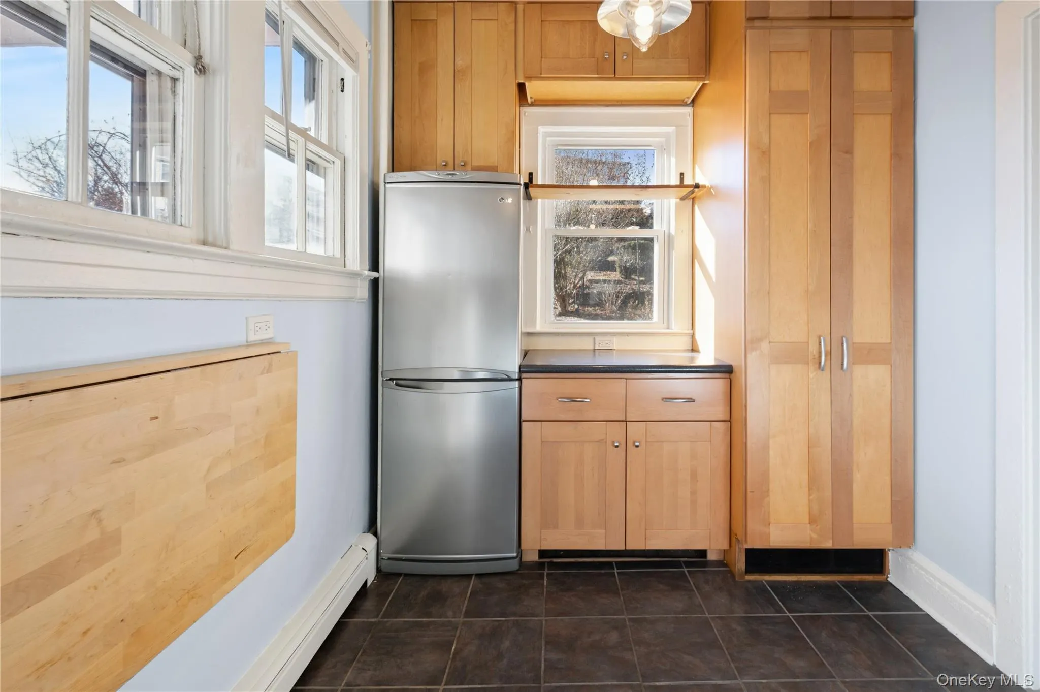 Kitchen featuring freestanding refrigerator, a baseboard heating unit, dark countertops, light brown cabinets, and dark tile patterned floors Kitchen featuring freestanding refrigerator, a baseboard heating unit, dark countertops, light brown cabinets, and dark tile patterned floors