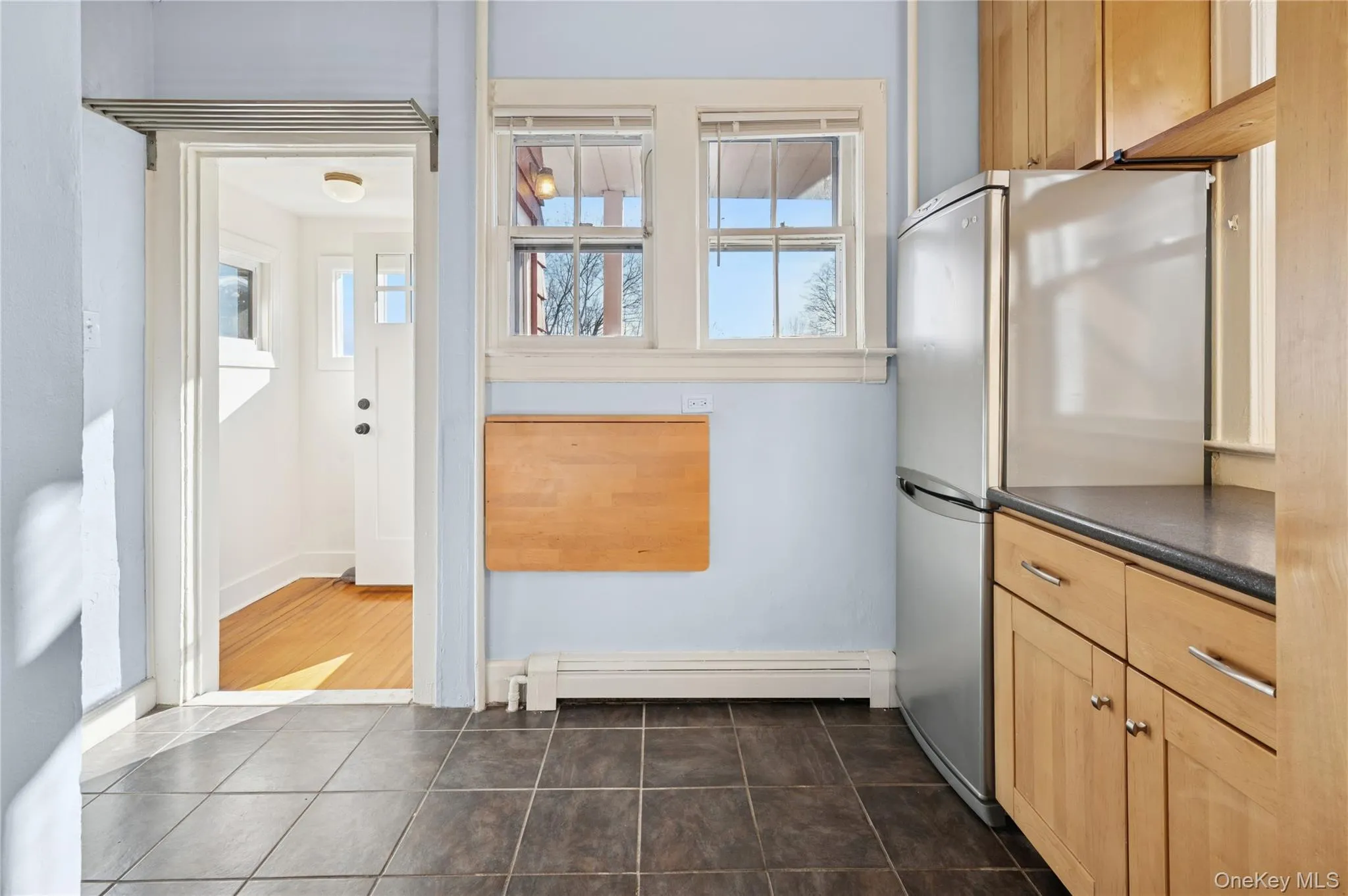 Kitchen with dark countertops, light brown cabinets, freestanding refrigerator, and a baseboard radiator Kitchen with dark countertops, light brown cabinets, freestanding refrigerator, and a baseboard radiator