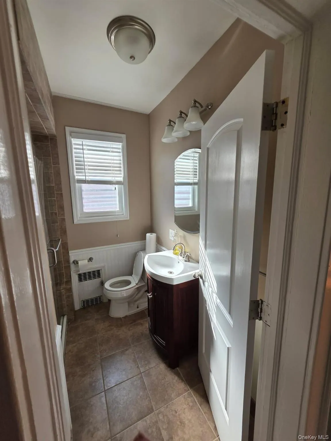 Full bathroom featuring vanity, a shower stall, light tile patterned flooring, radiator heating unit, and wainscoting Full bathroom featuring vanity, a shower stall, light tile patterned flooring, radiator heating unit, and wainscoting